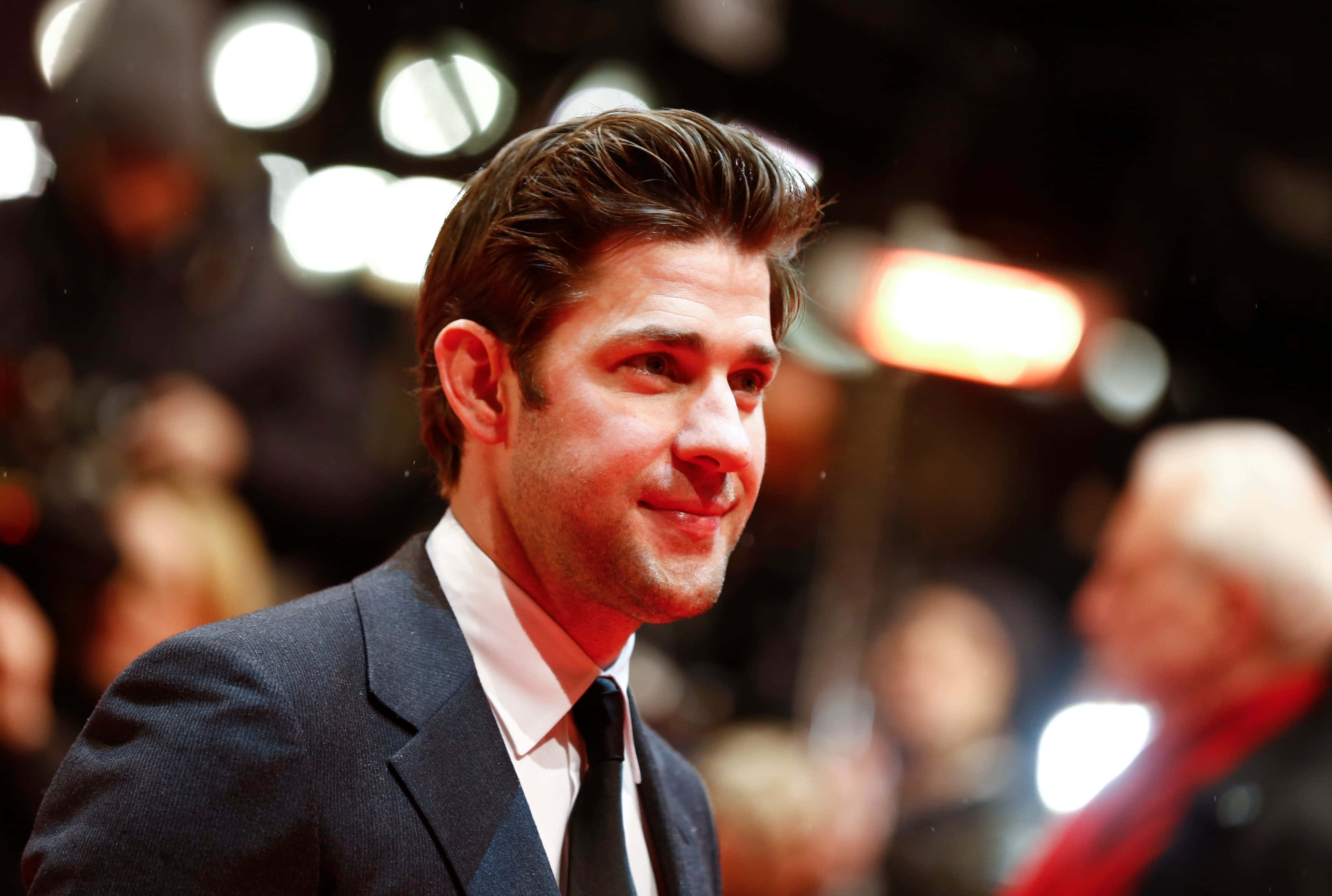 john krasinski 桌布 壁紙 — 一位身穿深色西裝的男子在明亮的活動燈光下溫暖地微笑，背景是模糊的人群。