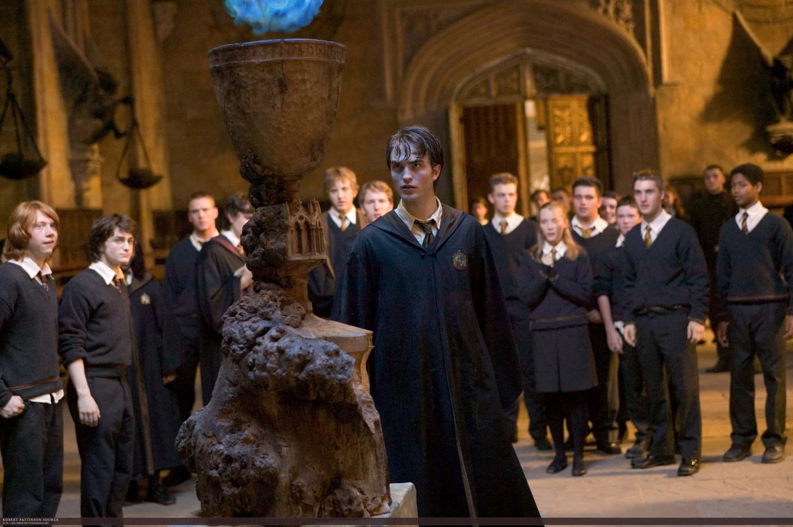 The Goblet Of Fire 桌布 壁紙 — 一位身穿深色長袍的年輕巫師站在一個宏偉的石廳中，面前是一個發光的藍色魔法高腳杯，周圍環繞著學生。