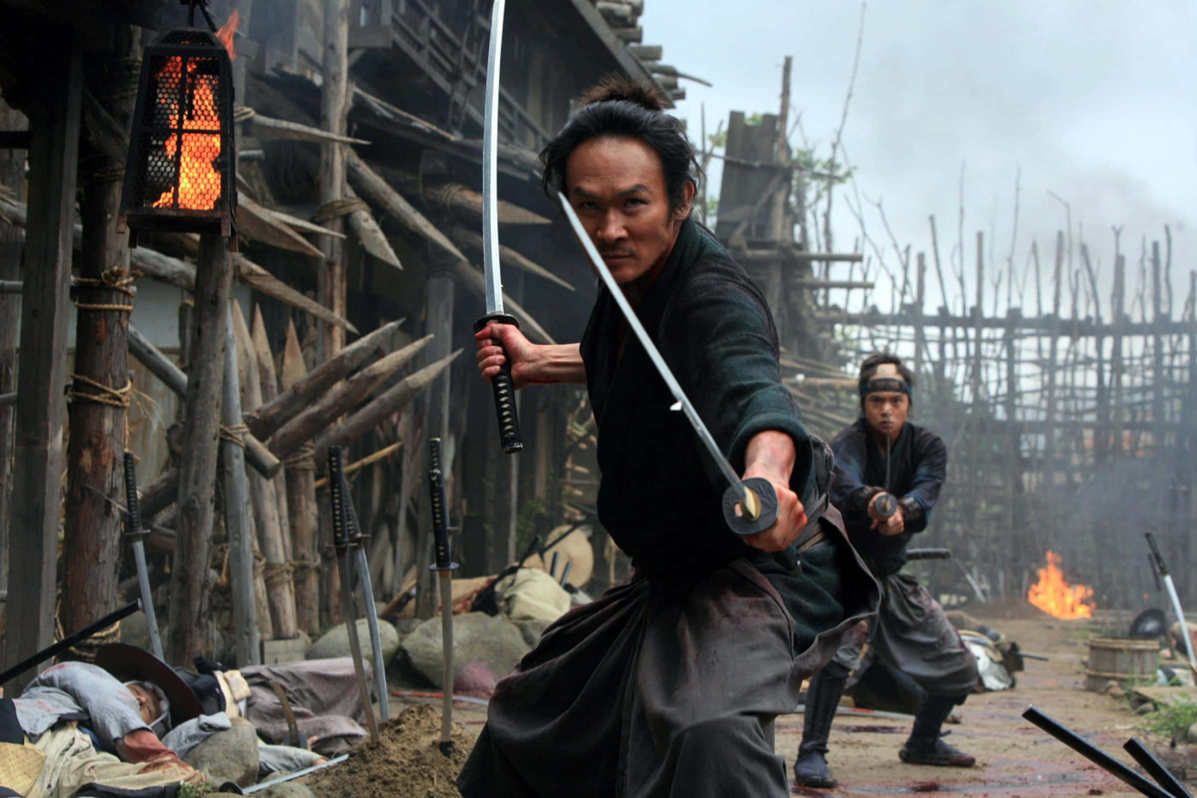Samurai Movies 桌布 壁紙 — 一位身穿深色長袍的武士武士手持武士刀在煙霧瀰漫的木村中向前衝刺，附近火焰燃燒，另一名戰士準備就緒。