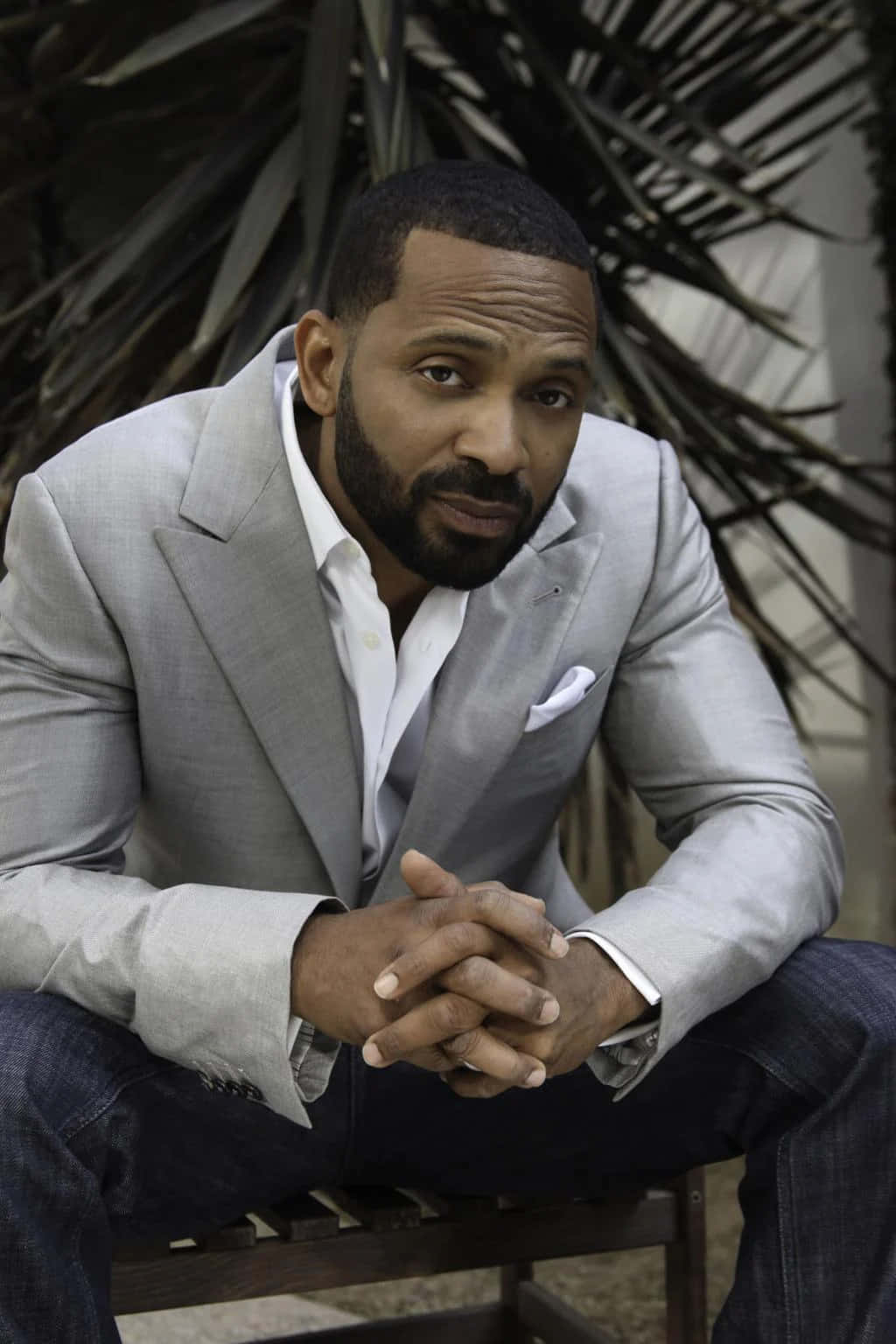 Mike Epps 桌布 壁紙 — 一位身穿淺灰色西裝的男子自信地坐在木長椅上，身後有深色棕櫚葉。