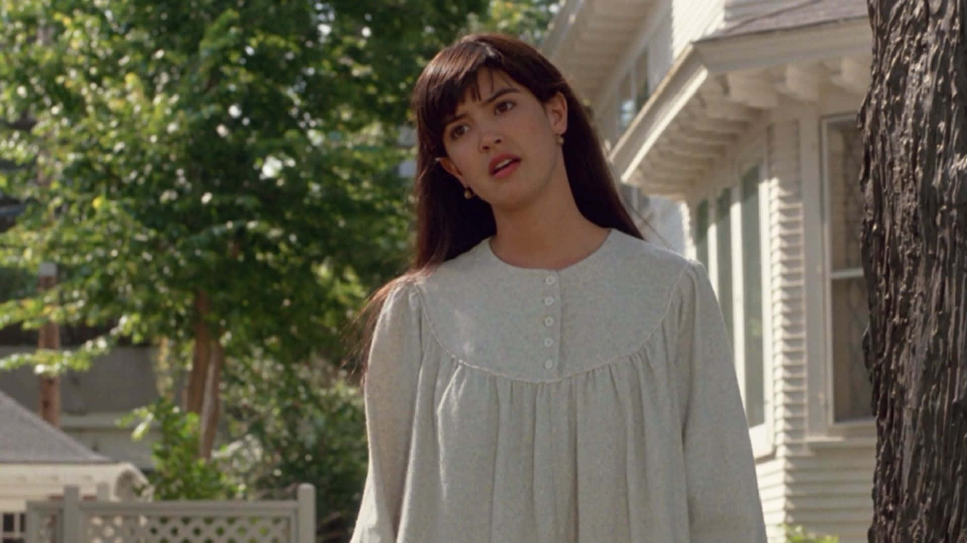 Phoebe Cates 桌布 壁紙 — 一位身穿淺灰色連衣裙的年輕女性站在戶外，靠近一棵樹和房子，表情驚訝。