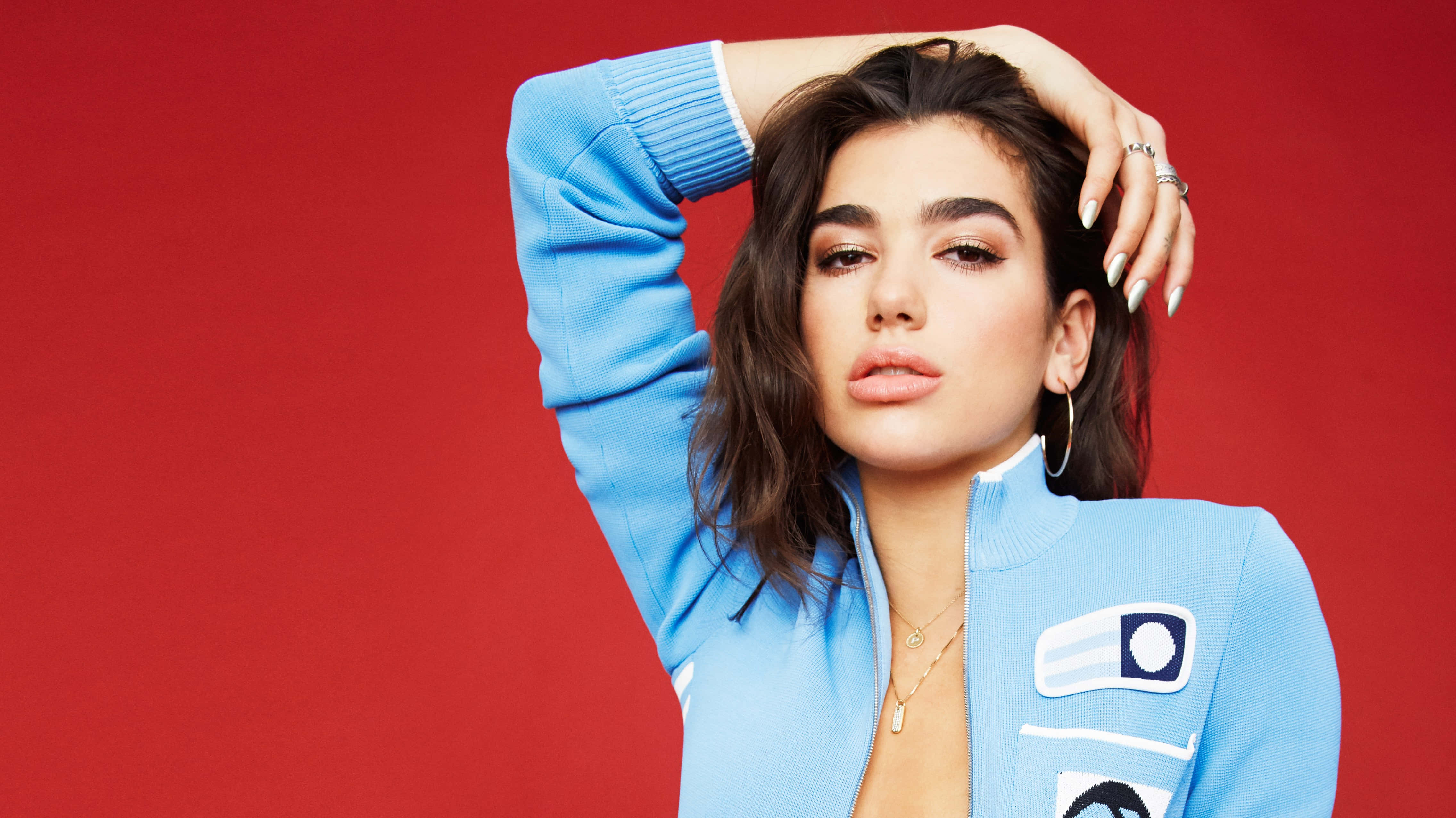 Dua Lipa 桌布 壁紙 — 一位身穿淺藍色外套的女性，在鮮豔的紅色背景前自信地擺姿勢，一手放在頭髮上。