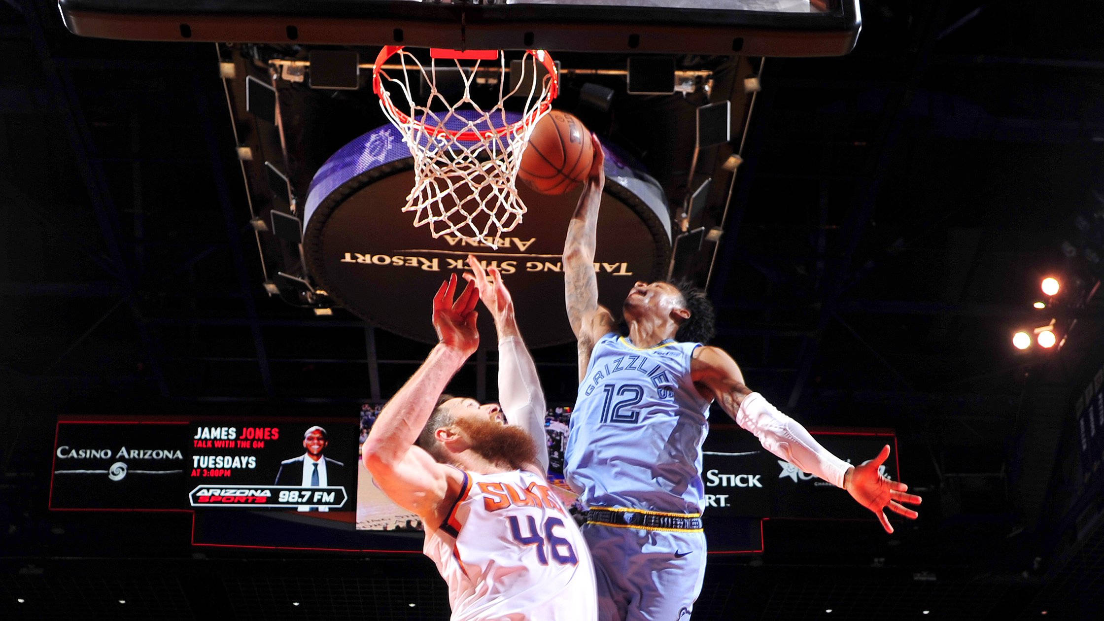Suns 桌布 壁紙 — 一位身穿淺藍色球衣的籃球運動員正在灌籃，而一位身穿白色的防守球員在場館燈光下試圖封阻。