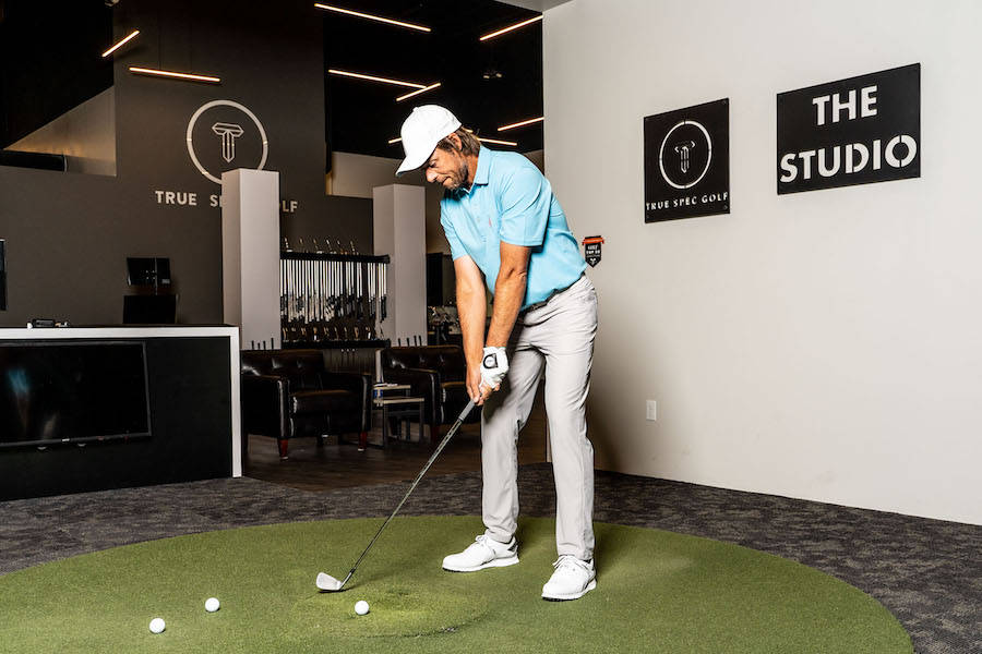 Aaron Baddeley True Spec Tour Team 壁紙 — 一位身穿淺藍色襯衫和白色帽子的高爾夫球手在一個帶有'THE STUDIO'標誌的綠色室內練習場揮桿。