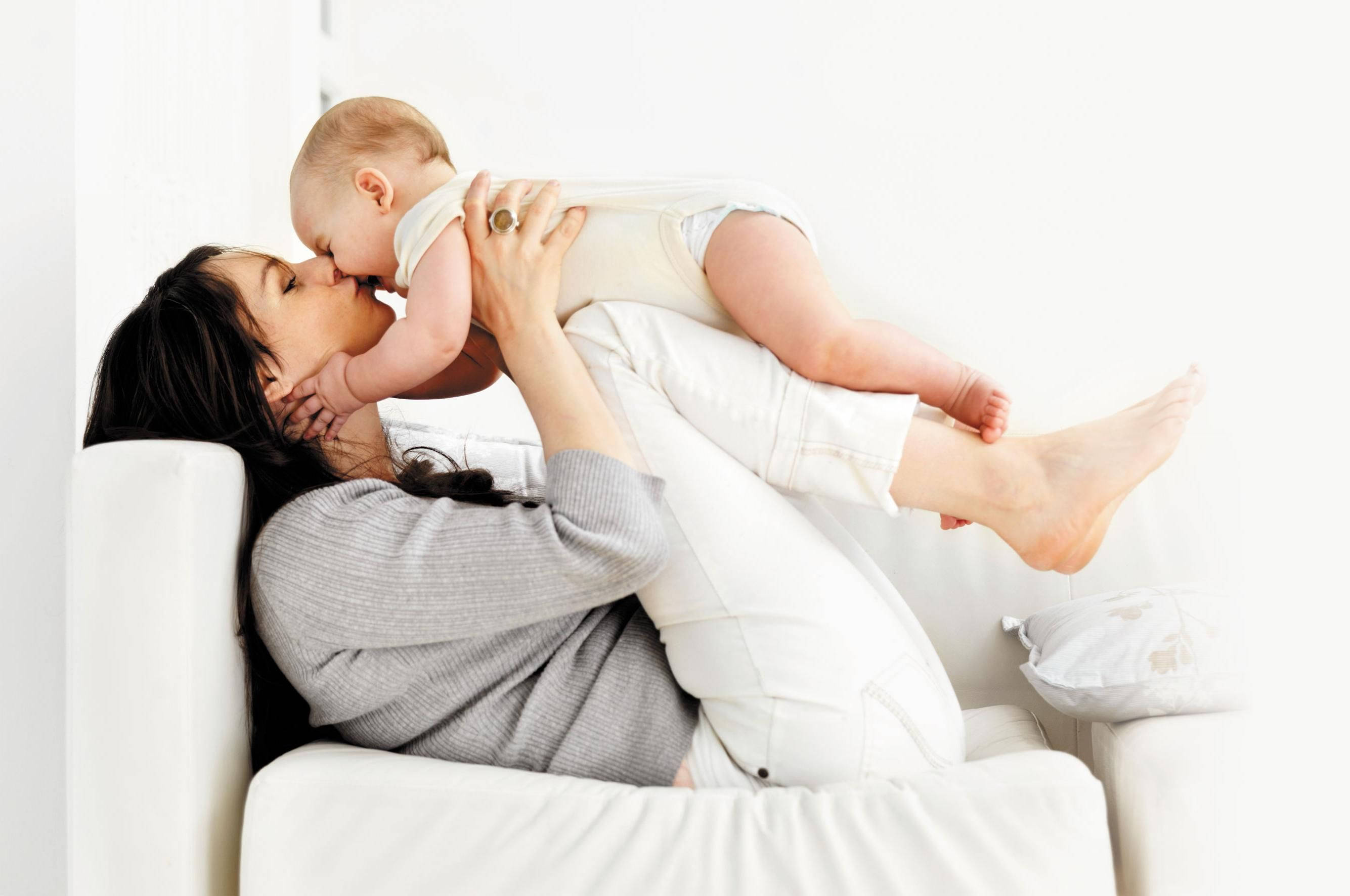 Mother And Baby 桌布 壁紙 — 一位身穿灰色毛衣的母親，坐在白色梳化上，親吻著她抱在懷裡的嬰兒。