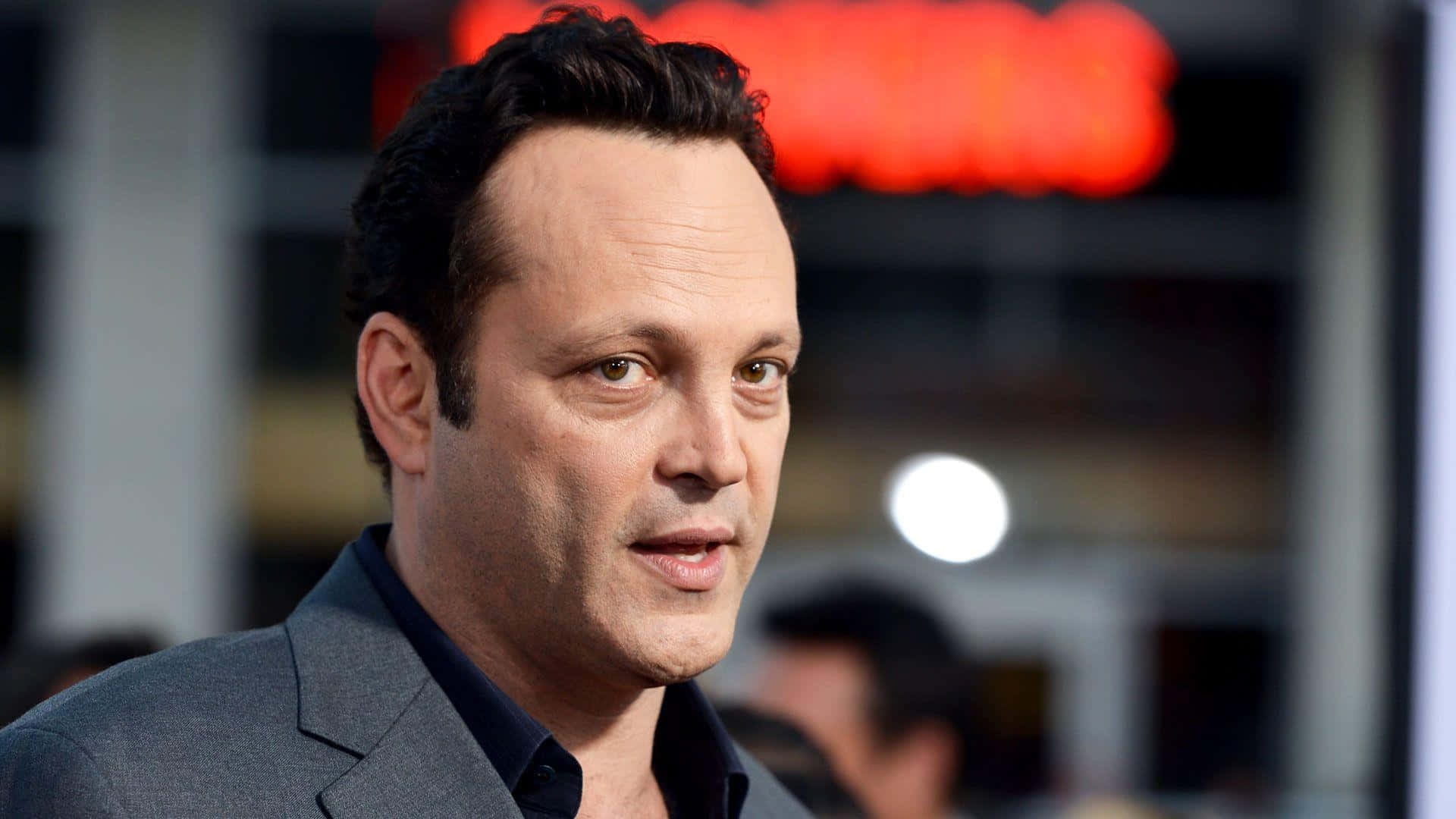 Vince Vaughn 桌布 壁紙 — 一位身穿灰色西裝的男士的特寫肖像，背景模糊並帶有紅色霓虹燈。