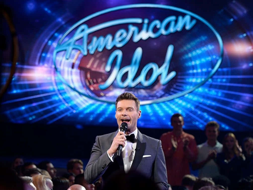 american idol 桌布 壁紙 — 一位身穿燕尾服的主持人在燈光明亮的美國偶像舞台上，對著咪高峰唱歌，背景有發光的藍色燈光和觀眾。