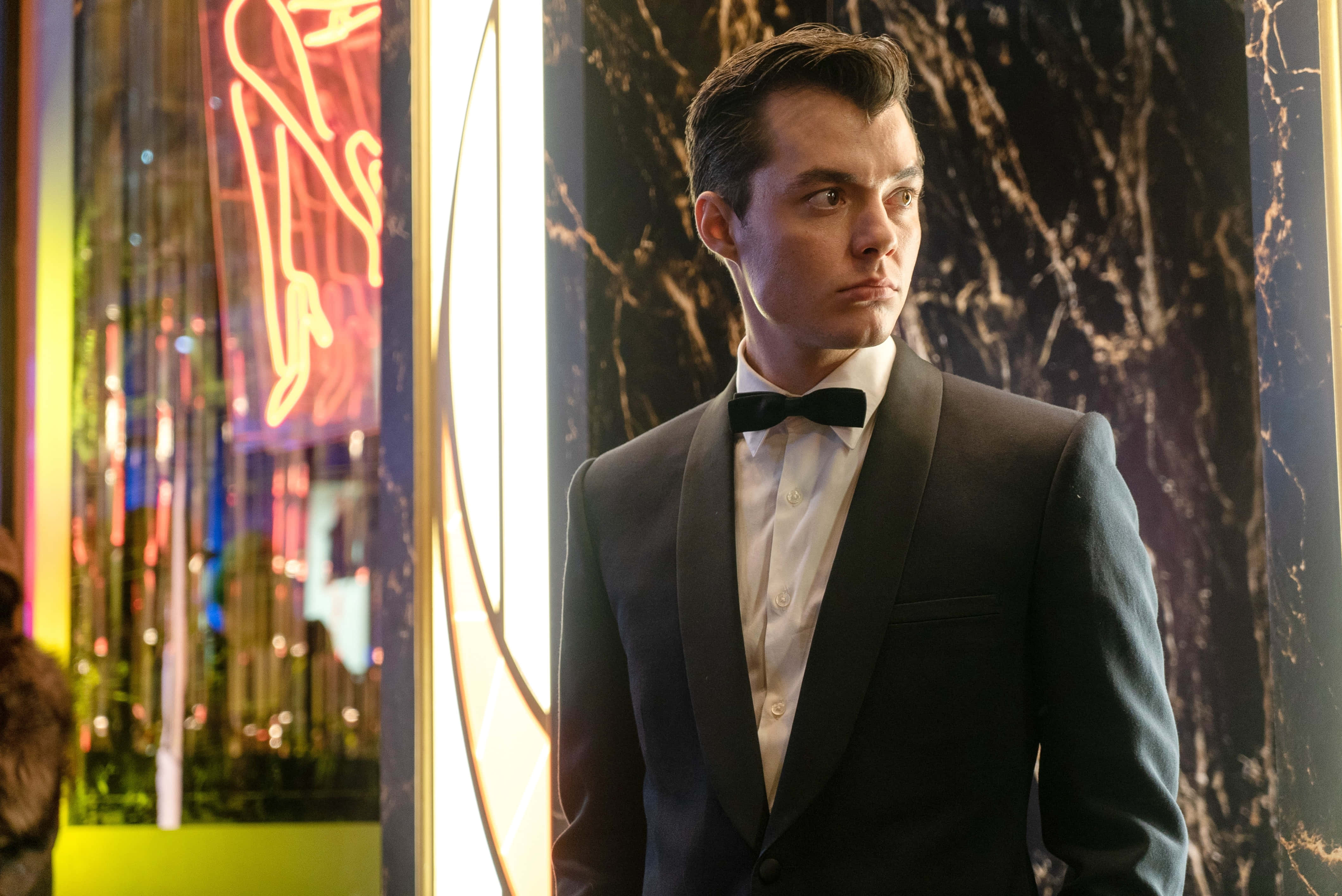 Alfred Pennyworth 桌布 壁紙 — 一位身穿燕尾服的男士，站在一個充滿霓虹燈和大理石牆壁的迷人夜總會中。