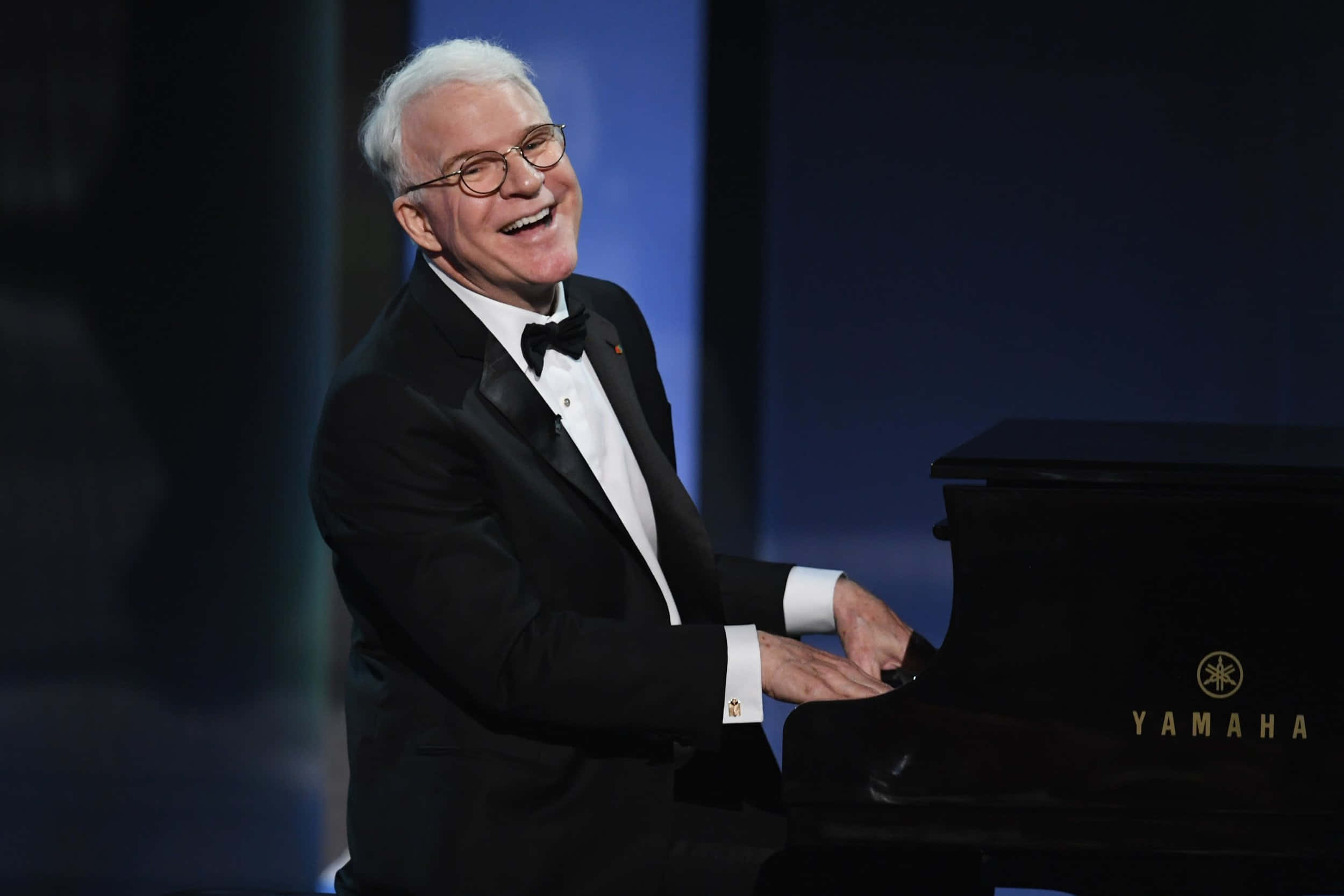 steve martin 桌布 壁紙 — 一位身穿燕尾服的較年長男士在燈光昏暗的舞台上愉快地笑著彈奏黑色Yamaha三角鋼琴。