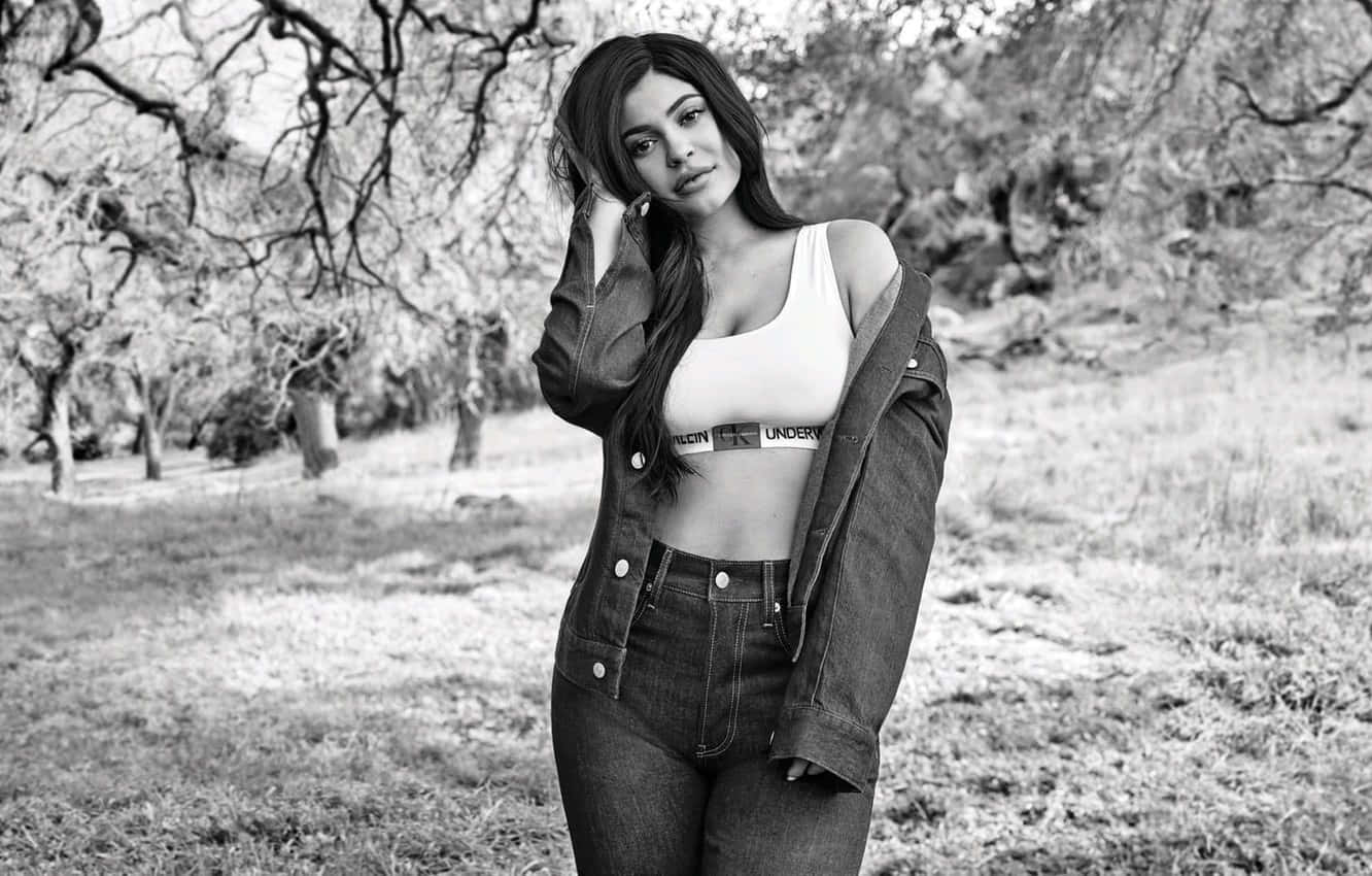 Black And White Celebrity 桌布 壁紙 — 一位身穿牛仔外套和牛仔褲的女性，在黑白照片中自信地站在無葉樹木和草地地形之間。