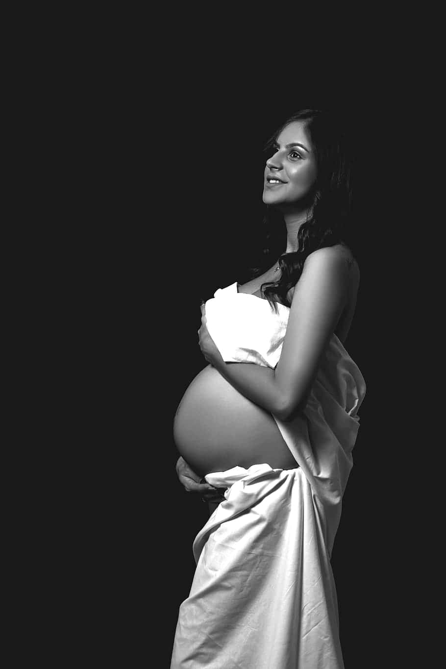 pregnancy 桌布 壁紙 — 一位身穿白布嘅孕婦喺純黑背景前輕輕微笑，抱住自己嘅肚。