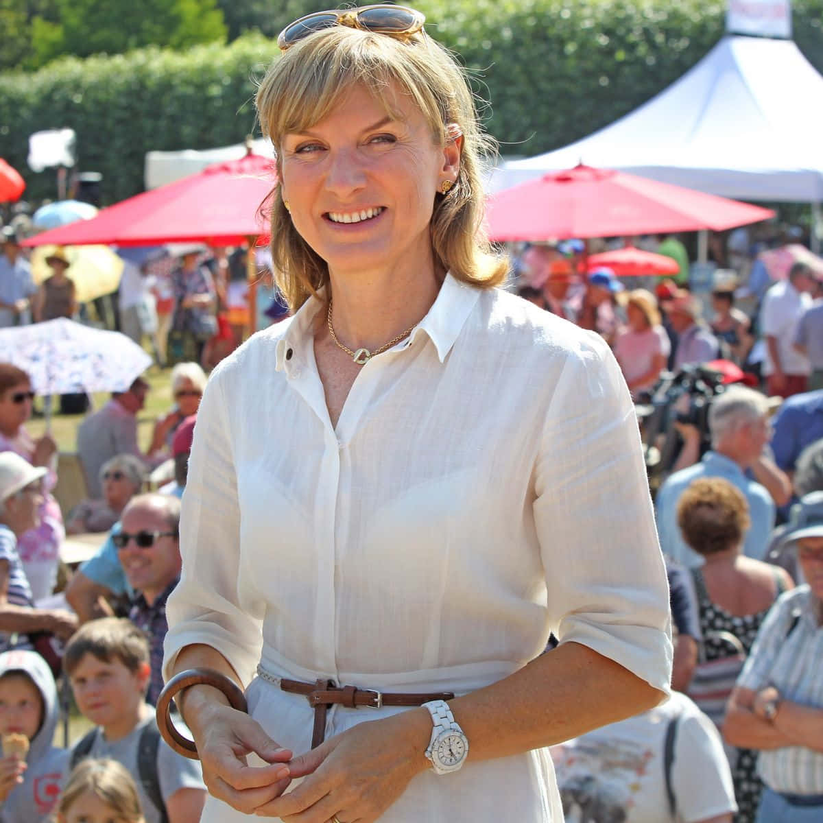 Fiona Bruce 桌布 壁紙 — 一位身穿白恤衫、面帶微笑的女性，站在陽光明媚的戶外市場，身後有紅色遮陽傘和人群。
