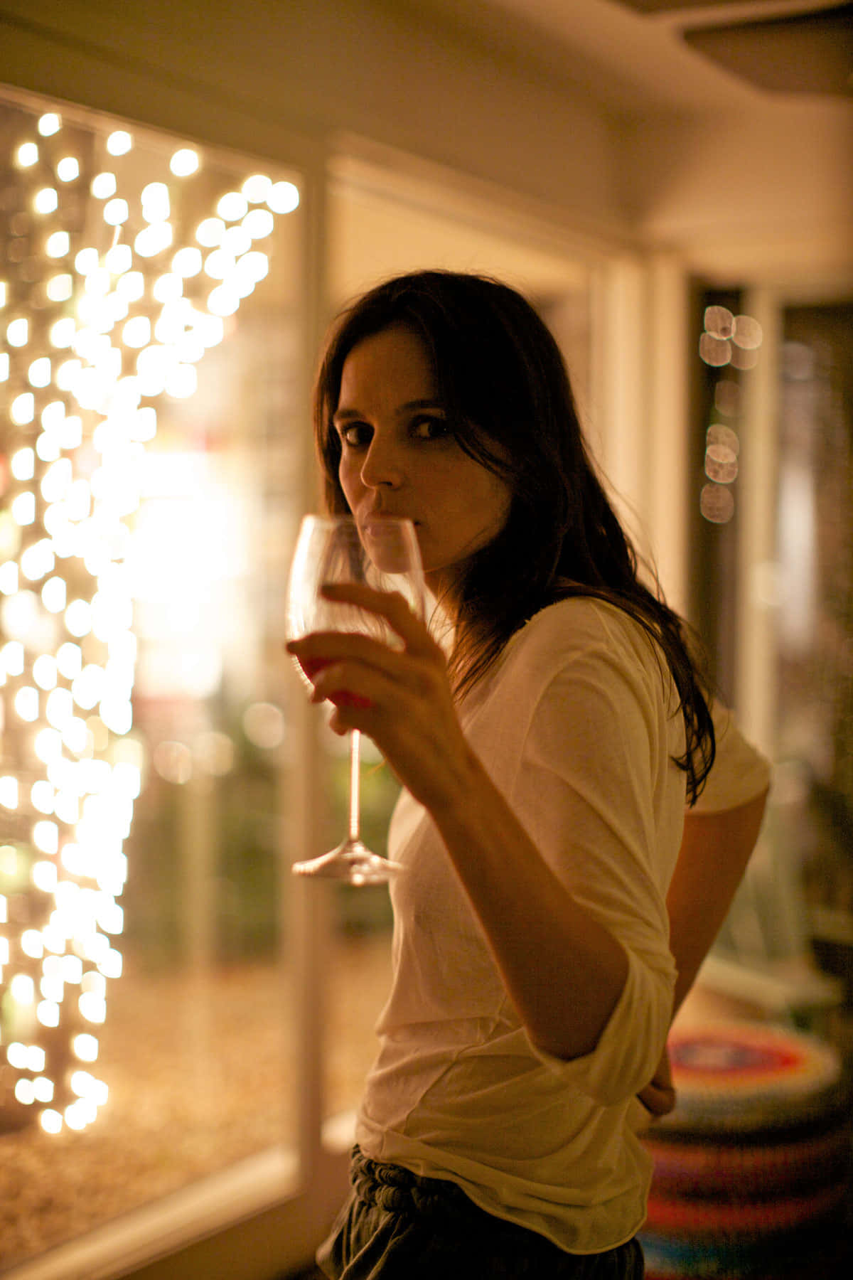 Elena Anaya 桌布 壁紙 — 一位身穿白恤衫的女性在室內啜飲葡萄酒，溫暖的串燈在她身後柔和地發光，營造出舒適的夜晚氛圍。