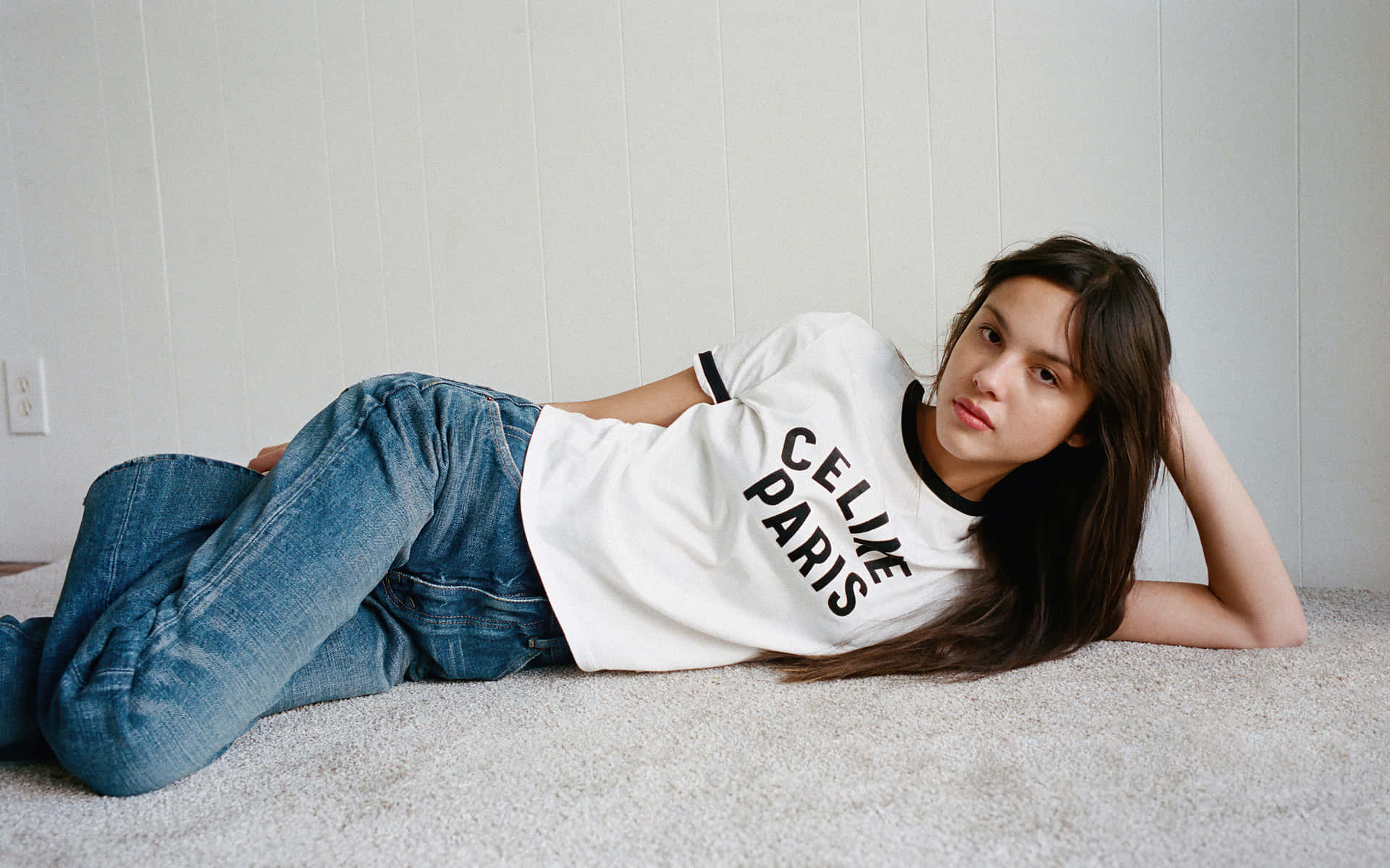 Olivia Rodrigo 桌布 壁紙 — 一位身穿白色 'CELINE PARIS' T恤和藍色牛仔褲的年輕女性，在淺色木鑲板牆邊的地毯上休息。