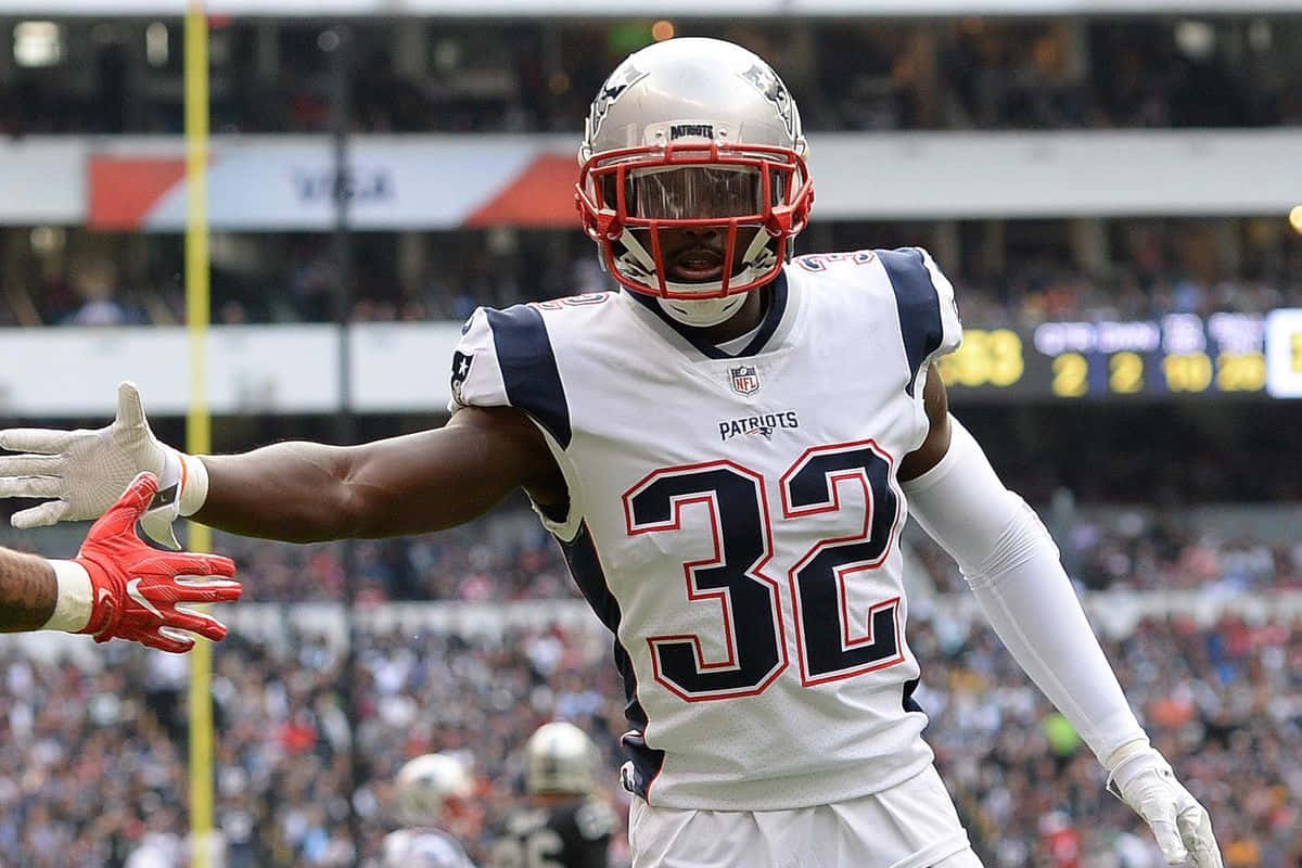 Devin Mccourty 桌布 壁紙 — 一位身穿白色32號球衣的新英格蘭愛國者隊美式足球員，在陽光明媚的球場上張開雙臂慶祝。