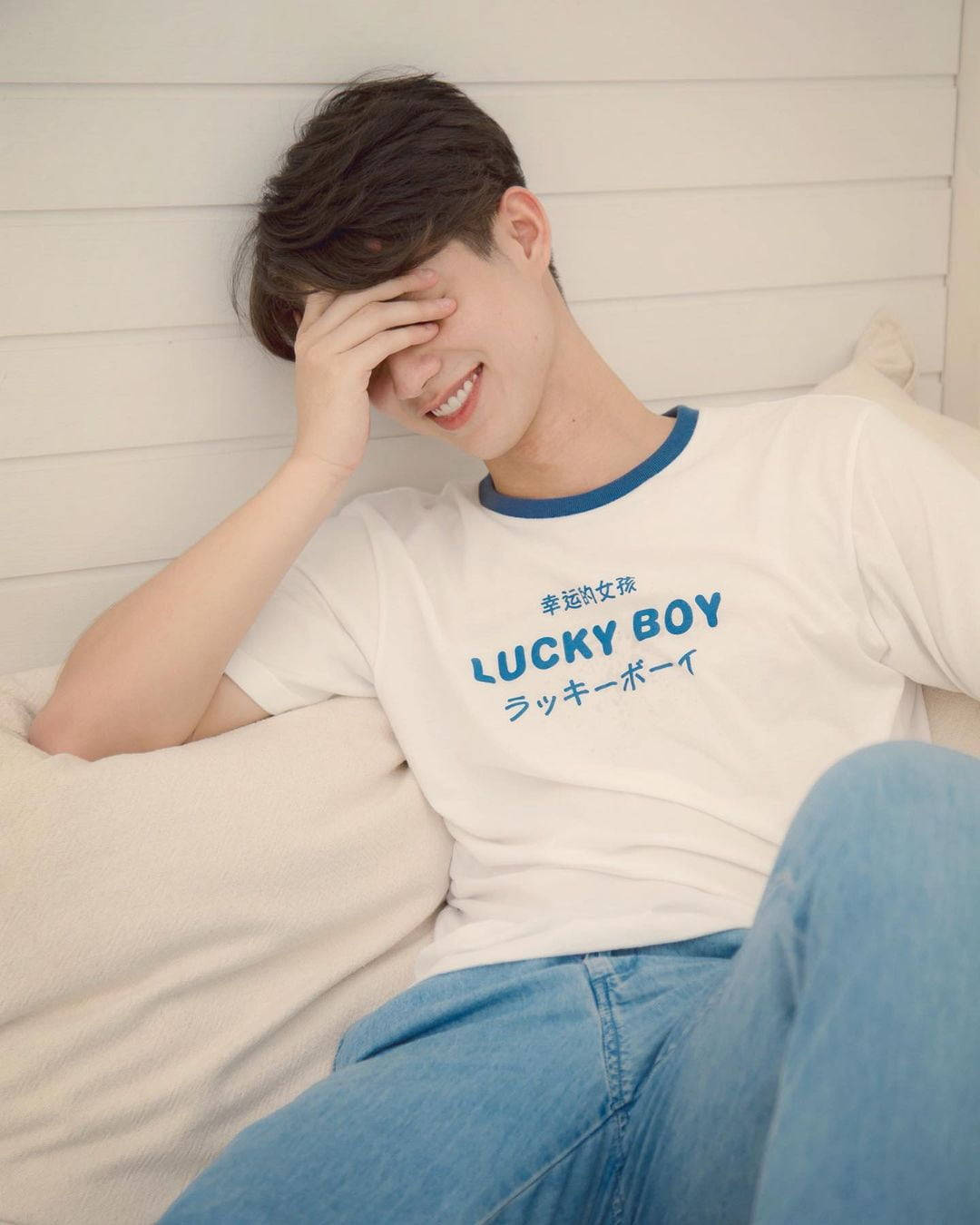 Bright Vachirawit 桌布 壁紙 — 一位身穿白色T恤、帶有藍色飾邊和「LUCKY BOY」字樣的年輕男子，將頭靠在手上，對著白色木牆微笑。