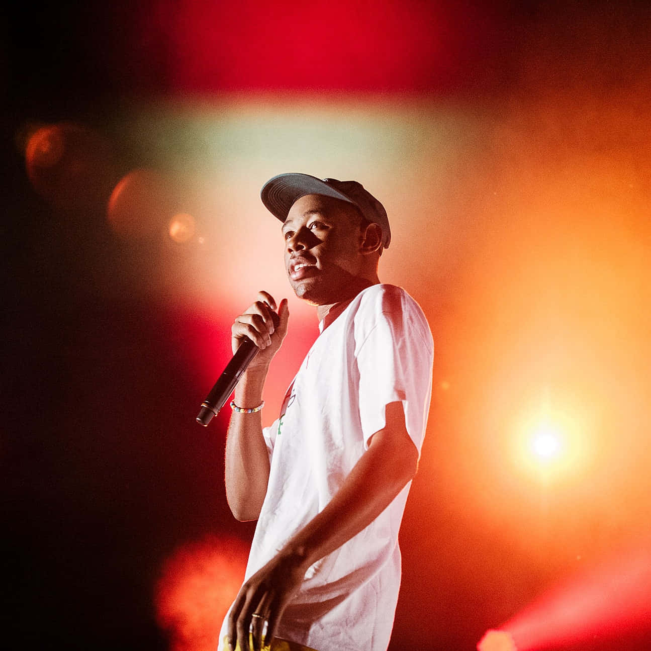 tyler the creator 桌布 壁紙 — 一位身穿白色T恤和帽子的男性表演者在充滿活力的紅色和橙色舞台燈光下對著麥克風唱歌。