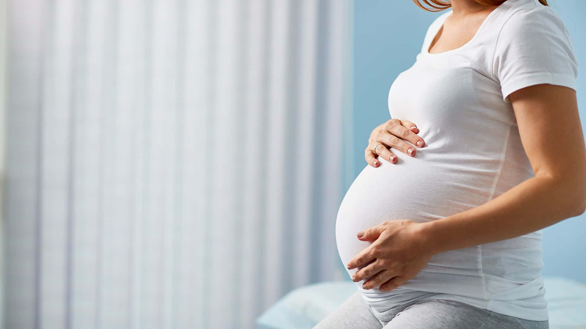 pregnancy 桌布 壁紙 — 一位身穿白色T恤的孕婦，在柔和的藍白背景下，用雙手輕輕地捧著腹部。