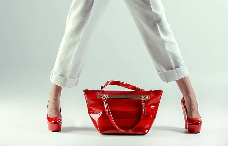 Red Handbag 桌布 壁紙 — 一位身穿白色九分褲和紅色漆皮高跟鞋的女性雙腿分開站立，在純白背景上，雙腳之間放著一個相配的紅色手袋。