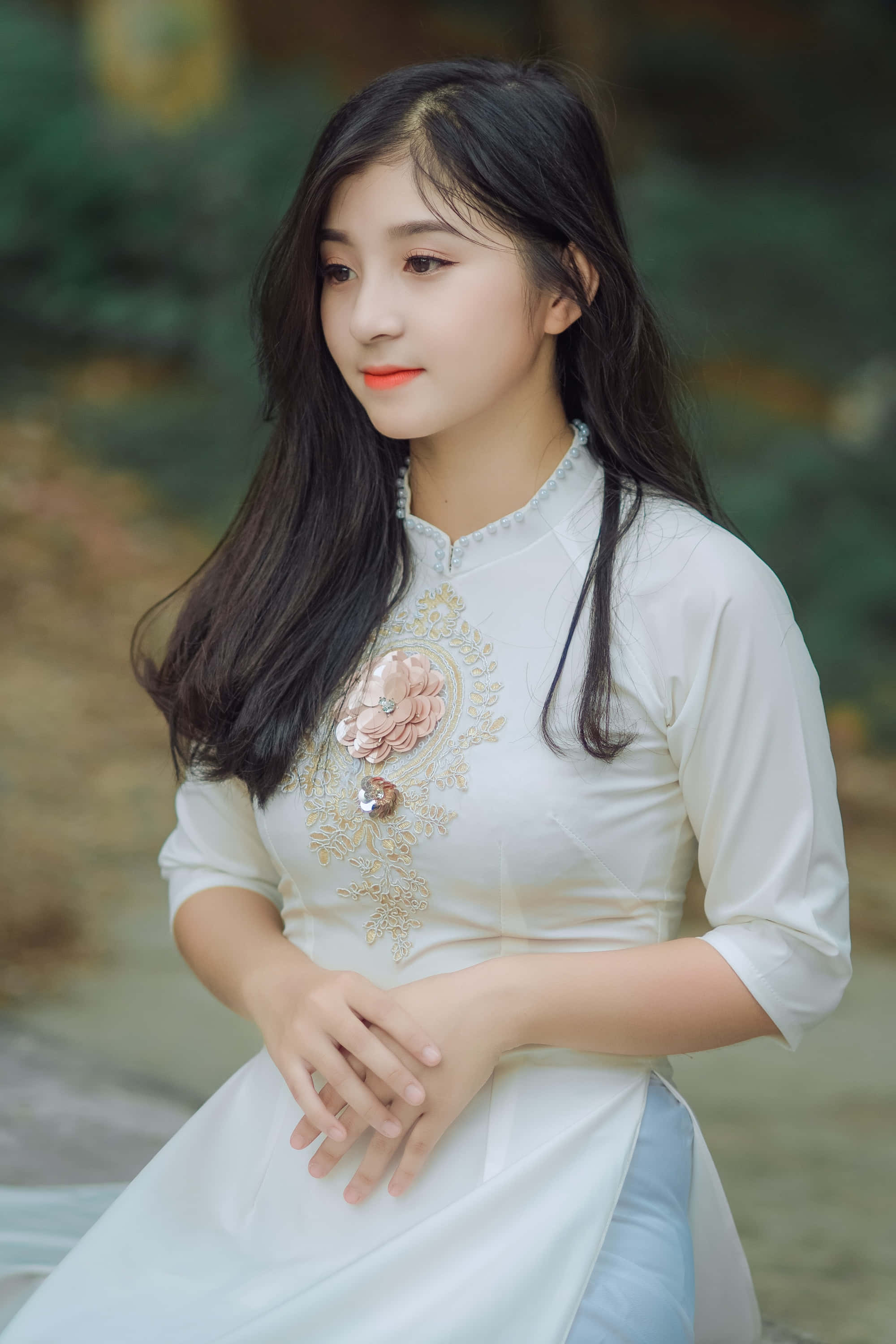 女孩 桌布 壁紙 — 一位身穿白色傳統刺繡花卉連衣裙的年輕女子，優雅地坐在柔和模糊的自然環境中。