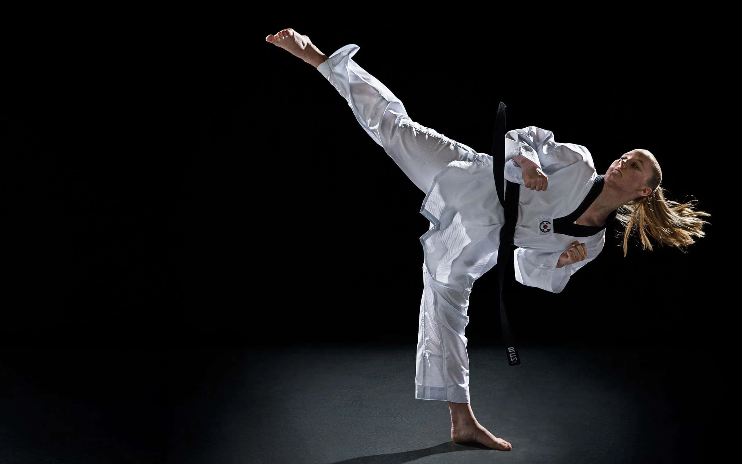 Martial Arts Uniform 桌布 壁紙 — 一位身穿白色制服、繫著黑色腰帶的女武術家，在純黑背景下執行高踢腿動作。