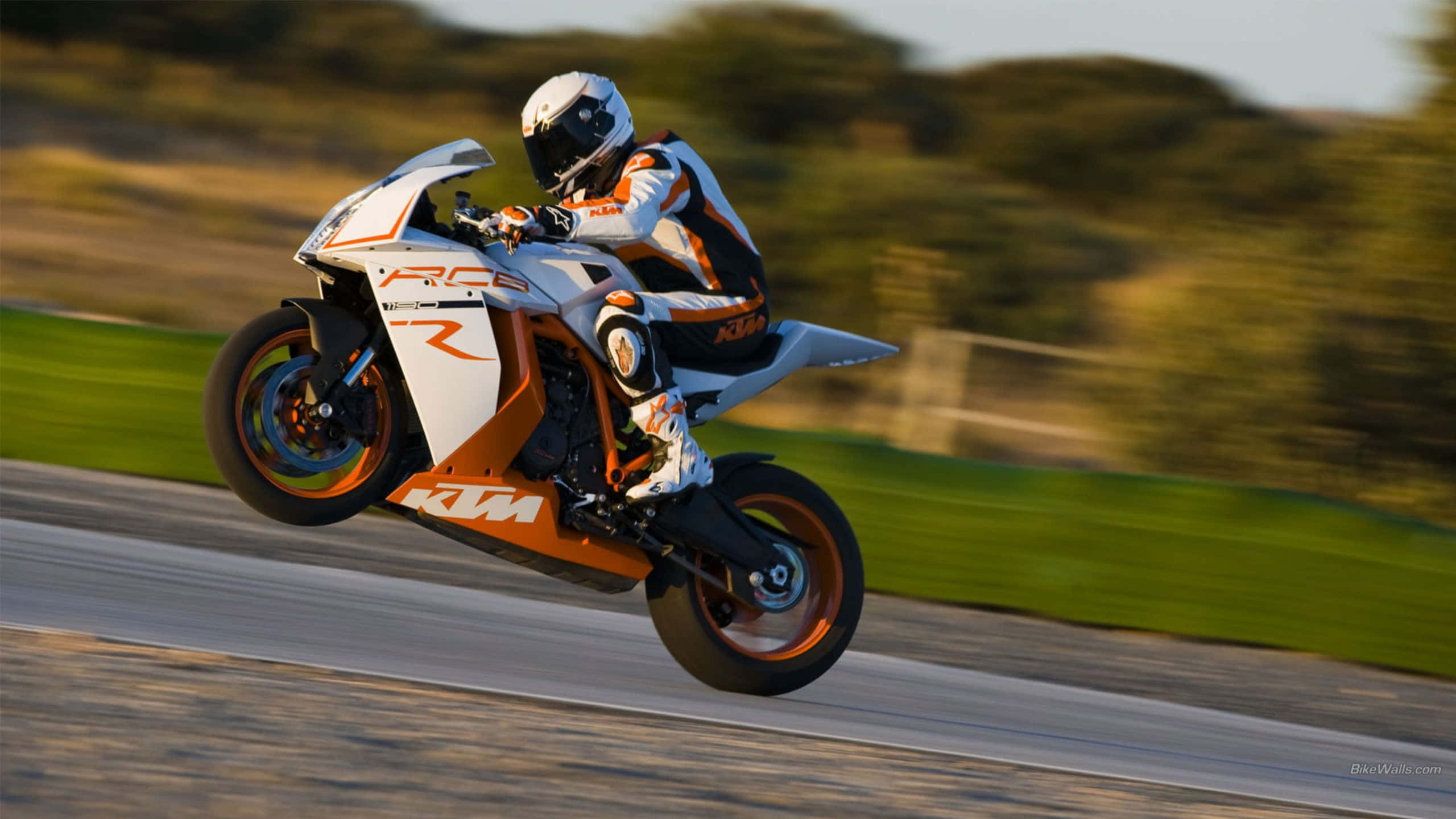 Ktm Bike 桌布 壁紙 — 一位身穿白色和橙色裝備的騎手，在陽光明媚的賽道上，騎著白色和橙色的KTM RC8R電單車傾斜入彎，背景是模糊的綠色植物。