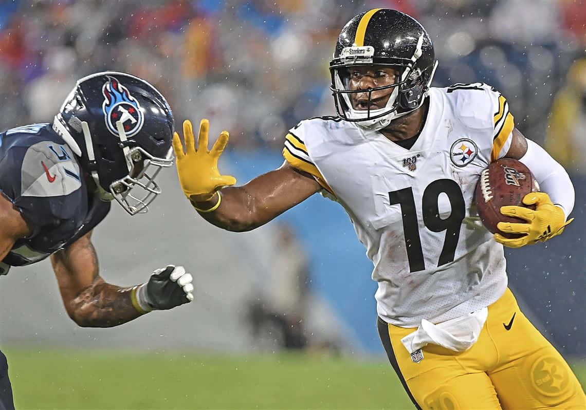 Juju Smith Schuster 桌布 壁紙 — 一位身穿白色和黃色球衣的匹茲堡鋼人隊球員帶著美式足球奔跑，而田納西泰坦隊的防守球員在潮濕的場地上飛身擒抱他。