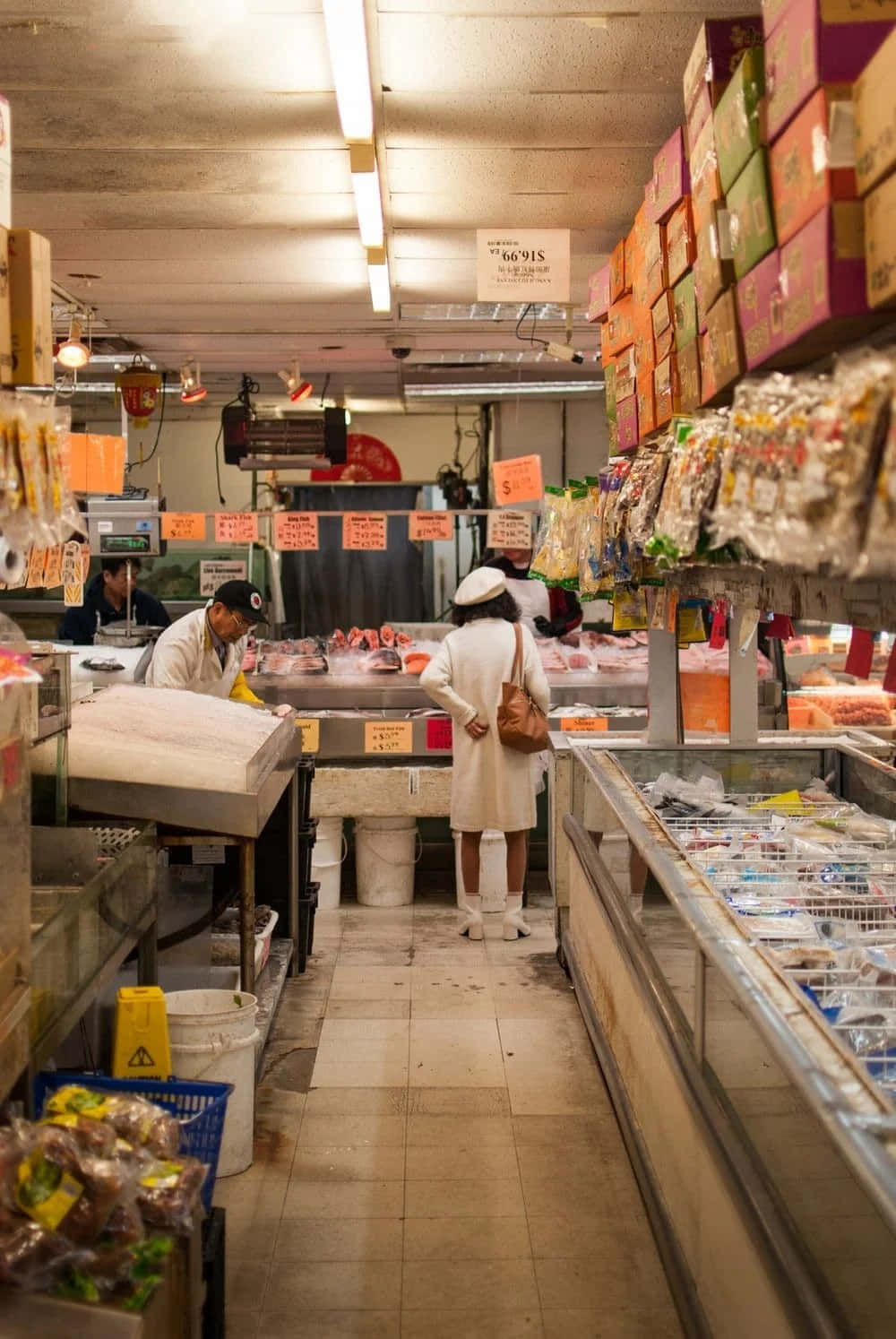 supermarket 桌布 壁紙 — 一位身穿白色外套的女性在繁忙的室內市場通道中瀏覽，通道兩旁擺放著冷藏海鮮和色彩繽紛的包裝商品，上方是螢光燈。