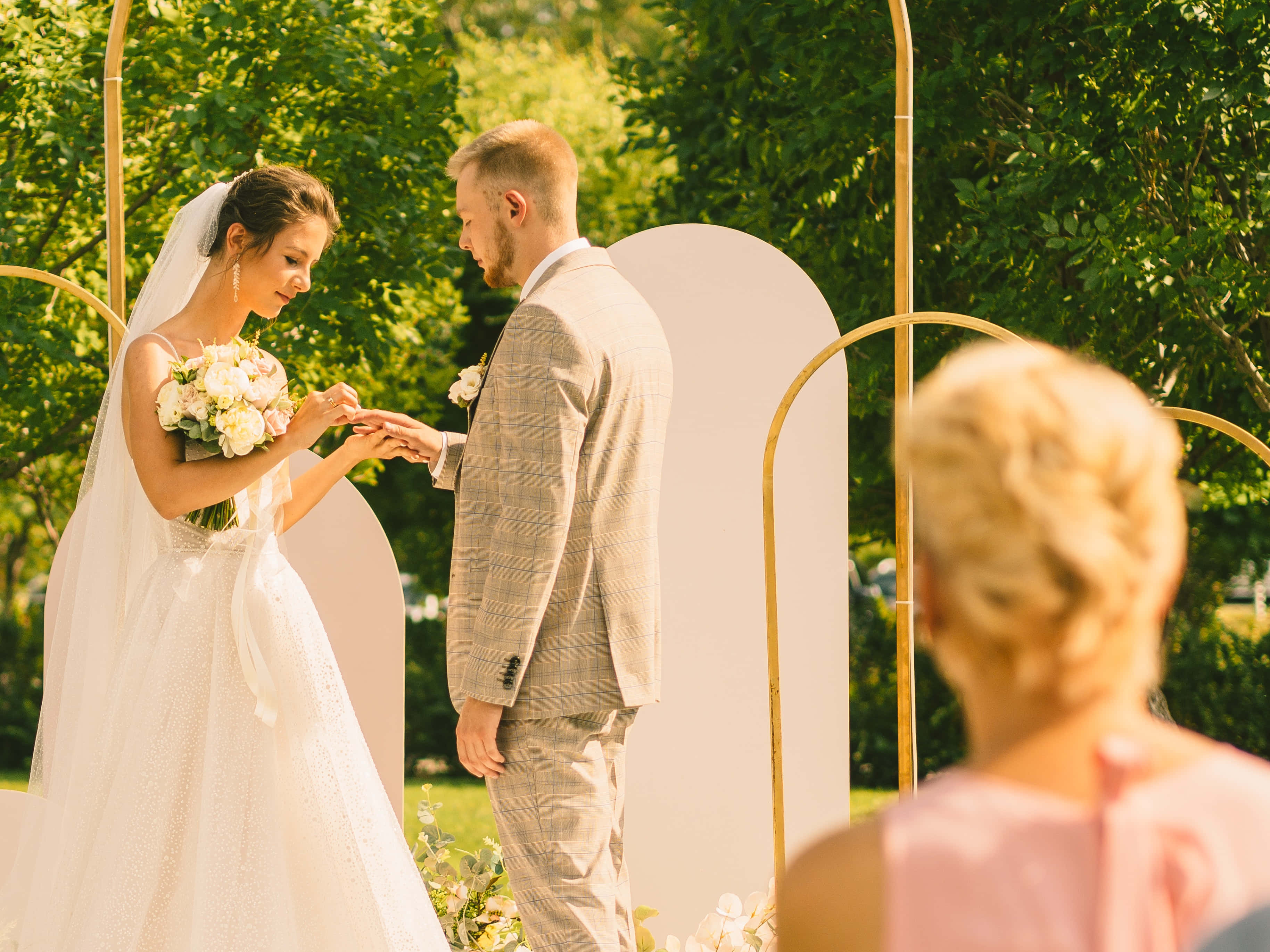Garden Wedding 桌布 壁紙 — 一位身穿白色婚紗和頭紗的新娘，在由茂密綠樹環繞的金色拱形婚禮拱門下，輕輕地將戒指戴在新郎的手指上。