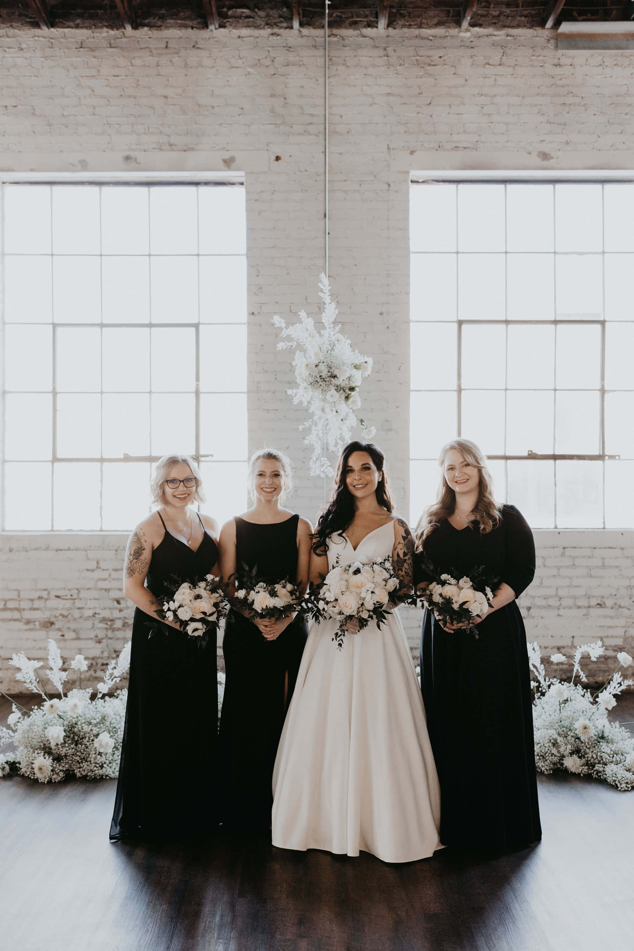 Black And White Wedding 桌布 壁紙 — 一位身穿白色婚紗的新娘與三位身穿黑色連衣裙的伴娘，手持花束，站在有大型窗戶的明亮磚牆前。