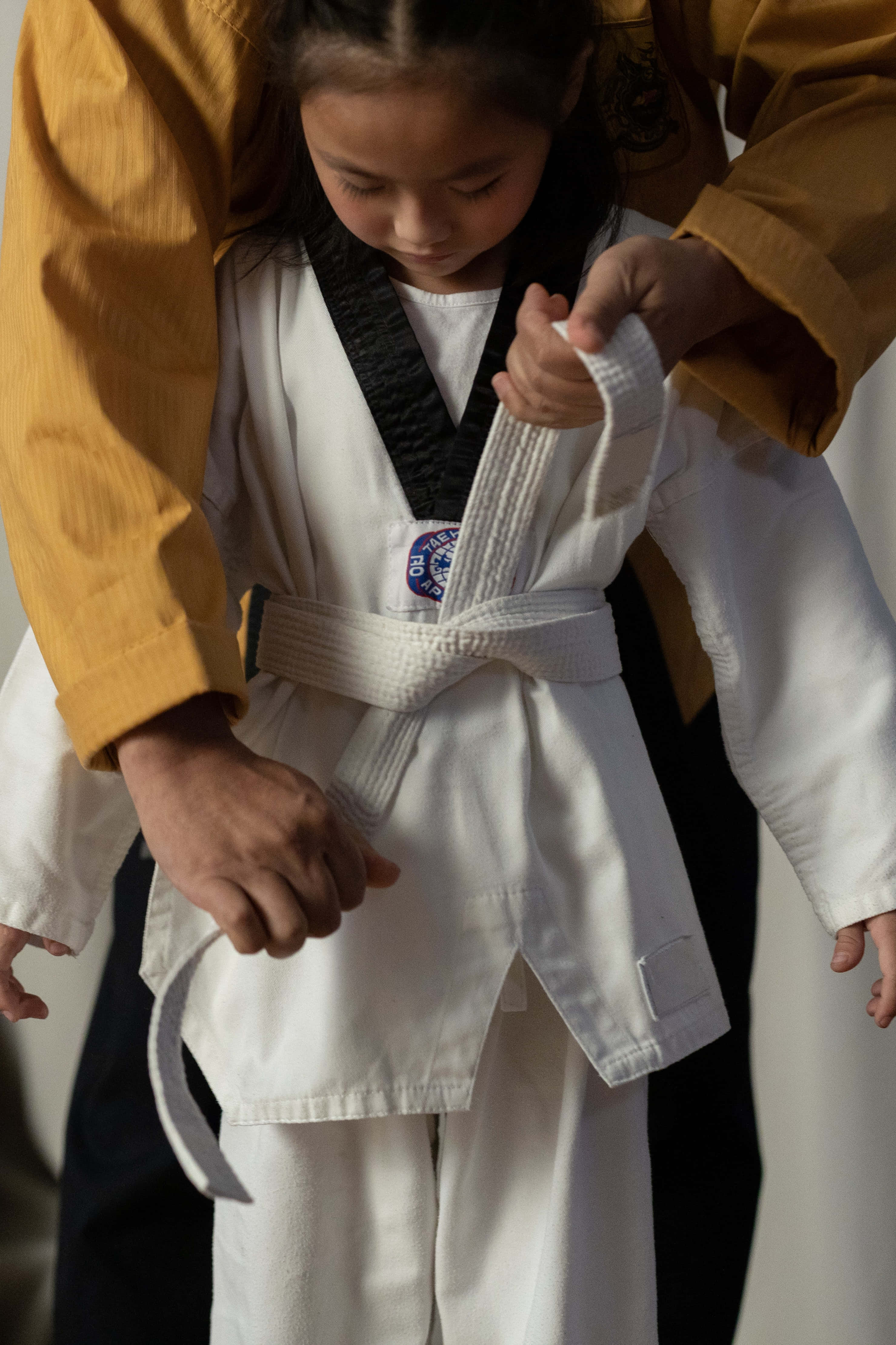 Taekwondo Uniform 桌布 壁紙 — 一位身穿白色武術服的小女孩，由一位身穿芥末黃外套的成年人為她繫上新的白色腰帶。