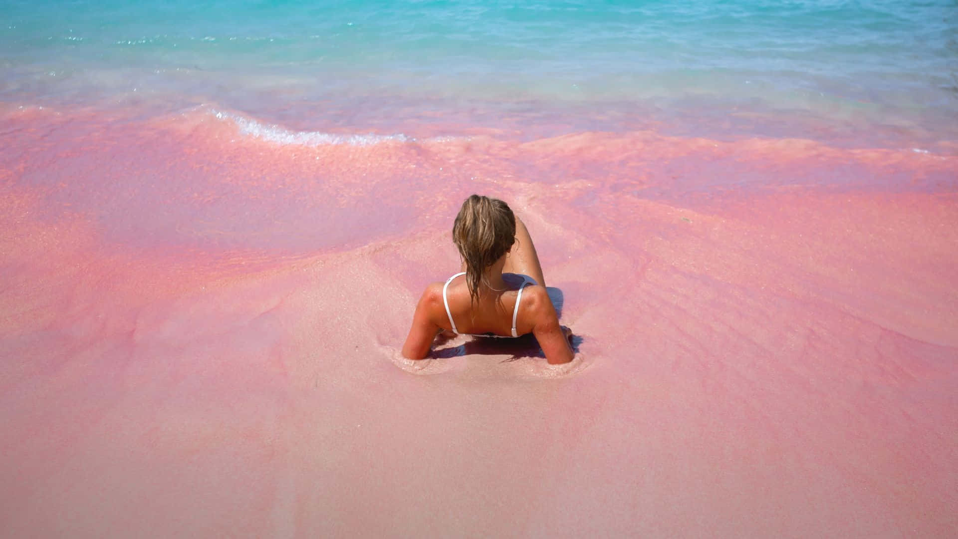 Pink Beach 桌布 壁紙 — 一位身穿白色比基尼的女性獨自坐在鮮豔的粉紅色沙灘上，在明亮的日光下，碧綠色的海浪輕輕拍打她的身旁。