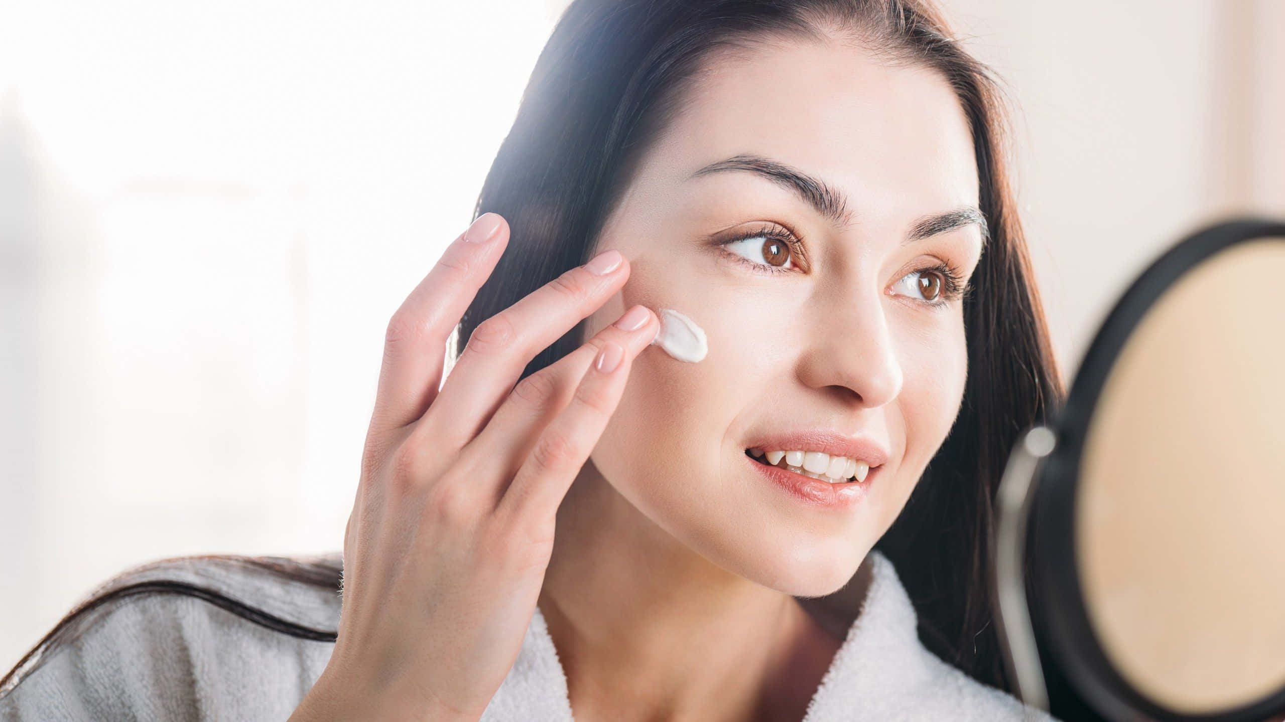 skin 桌布 壁紙 — 一位身穿白色浴袍的女性在圓鏡前微笑著將乳霜塗抹在臉頰上。