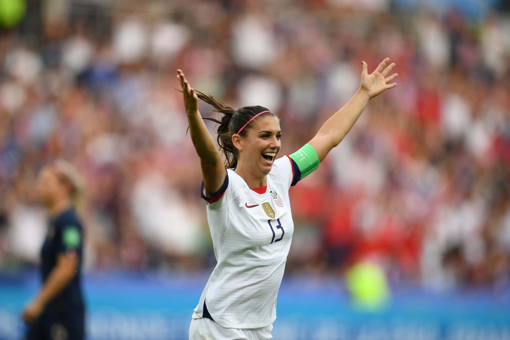 Alex Morgan 桌布 壁紙 — 一位身穿白色球衣的快樂女足球員在陽光普照的球場上高舉雙臂，露出燦爛笑容慶祝。
