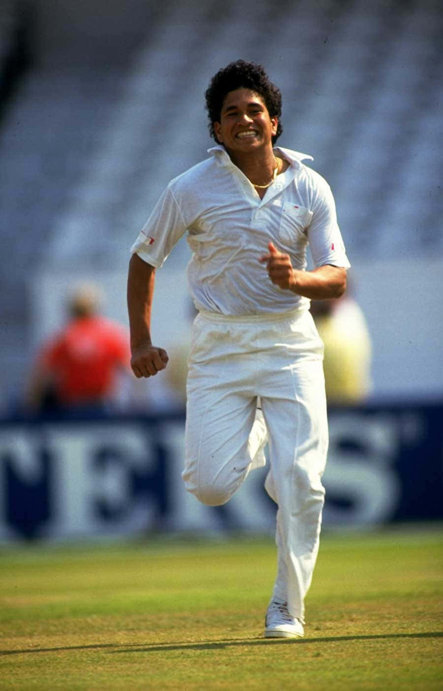 Sachin Tendulkar 桌布 壁紙 — 一位身穿白色球衣的快樂板球運動員在陽光明媚的綠色球場上奔跑，背景是模糊的體育場看台。