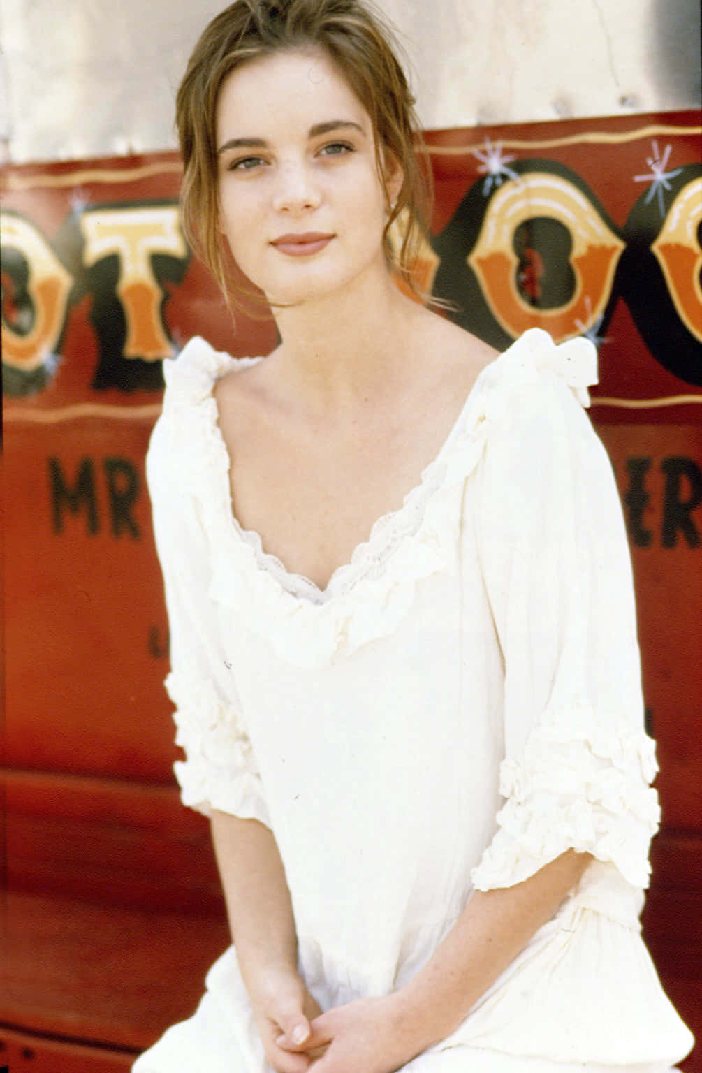 Gabrielle Anwar 桌布 壁紙 — 一位身穿白色荷葉邊連衣裙的年輕女子，平靜地坐在一輛帶有粗體字樣的復古紅色嘉年華馬車前。