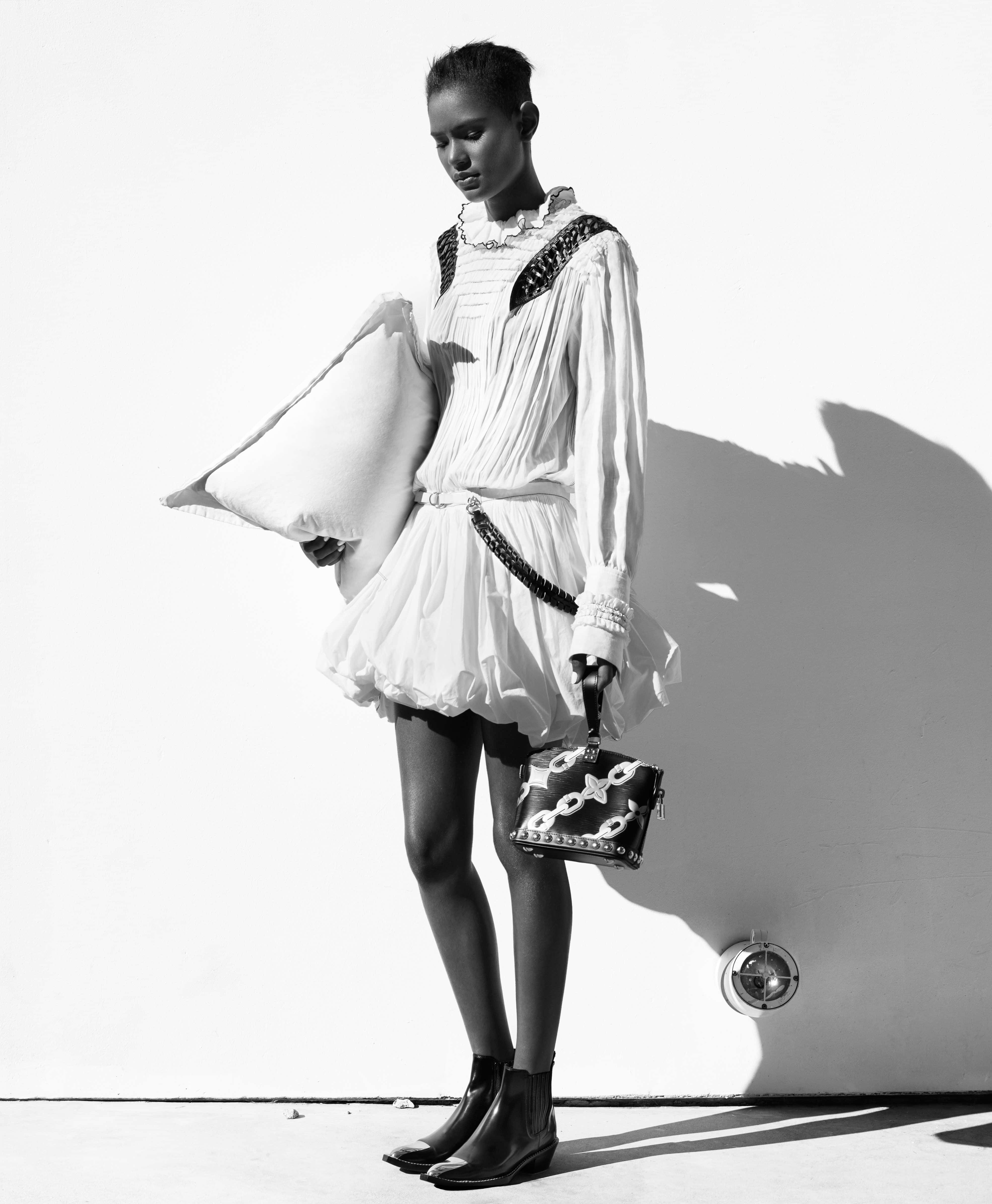 Black And White Fashion 桌布 壁紙 — 一位身穿白色荷葉邊連衣裙的模特兒，在純白牆壁前拿著枕頭和華麗手袋，投射出戲劇性的陰影。