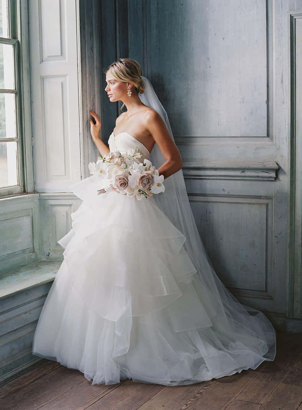 Bride's Dress 桌布 壁紙 — 一位身穿白色蓬鬆婚紗的新娘站在窗邊，手持一束白色和粉紅色的玫瑰，背景是風化的藍色牆壁。