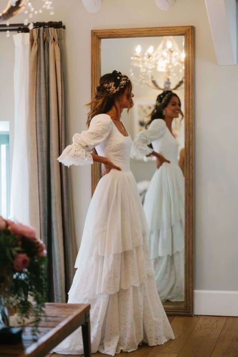 Wedding Dress 桌布 壁紙 — 一位身穿白色蕾絲婚紗、帶有鐘形袖的新娘，在溫暖照明的房間裡，欣賞著高木框鏡中的倒影。