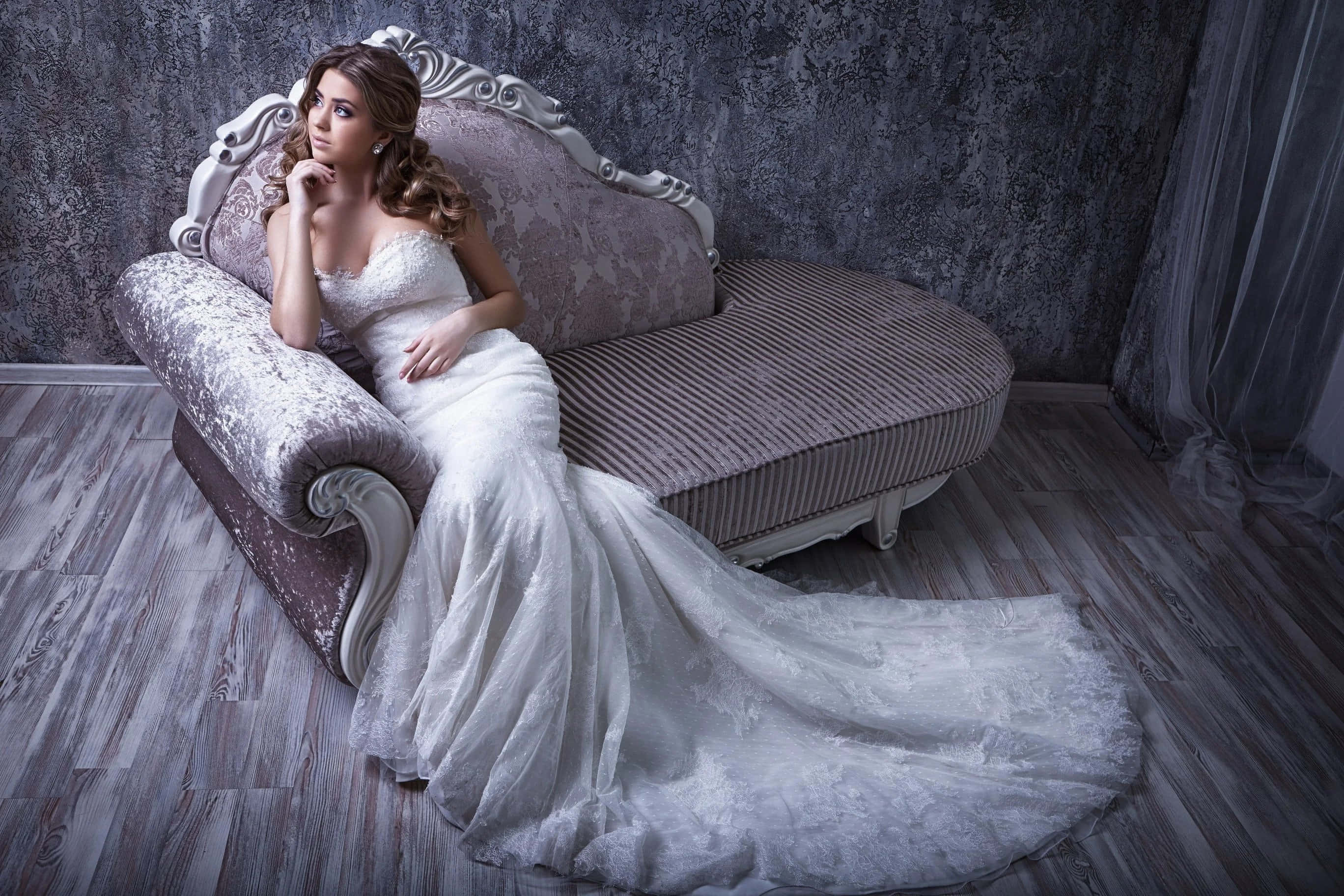 Wedding Dress 桌布 壁紙 — 一位身穿白色蕾絲禮服的新娘，在一個氣氛濃郁、帶有紋理的灰色房間裡，優雅地斜倚在一張復古貴妃椅上。
