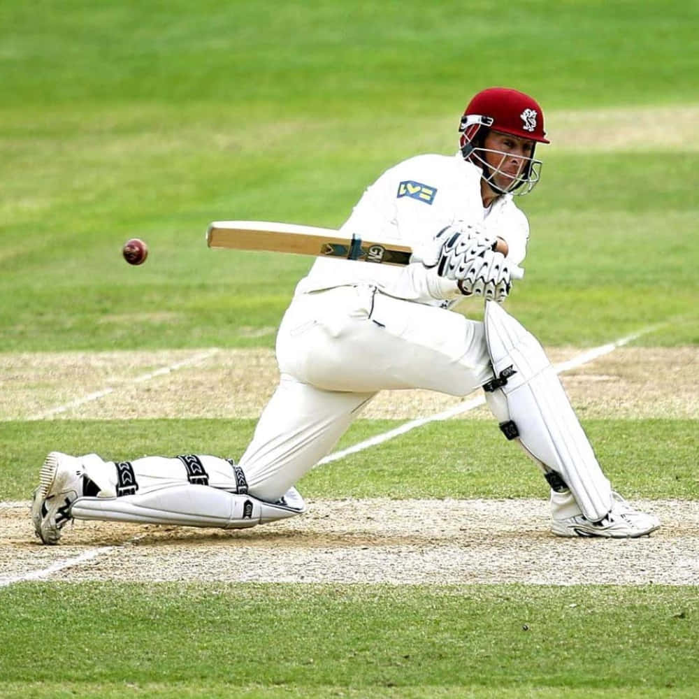 Cricket 桌布 壁紙 — 一位身穿白色裝備和紅色頭盔的板球擊球手在綠色場地上向一個紅色球揮動球棒。