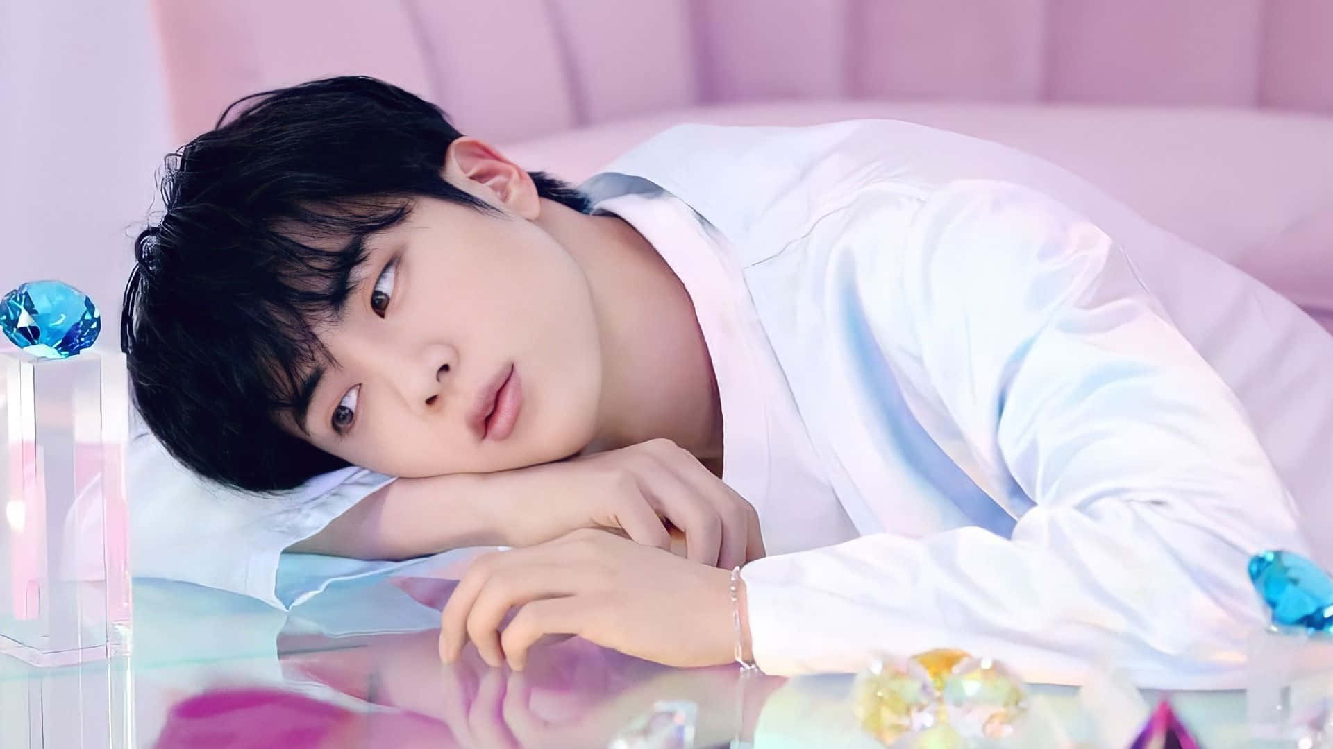 Jin 桌布 壁紙 — 一位身穿白色襯衫的年輕男子在反光表面上將頭靠在手臂上休息，周圍環繞著發光的藍色水晶和粉紅色背景。