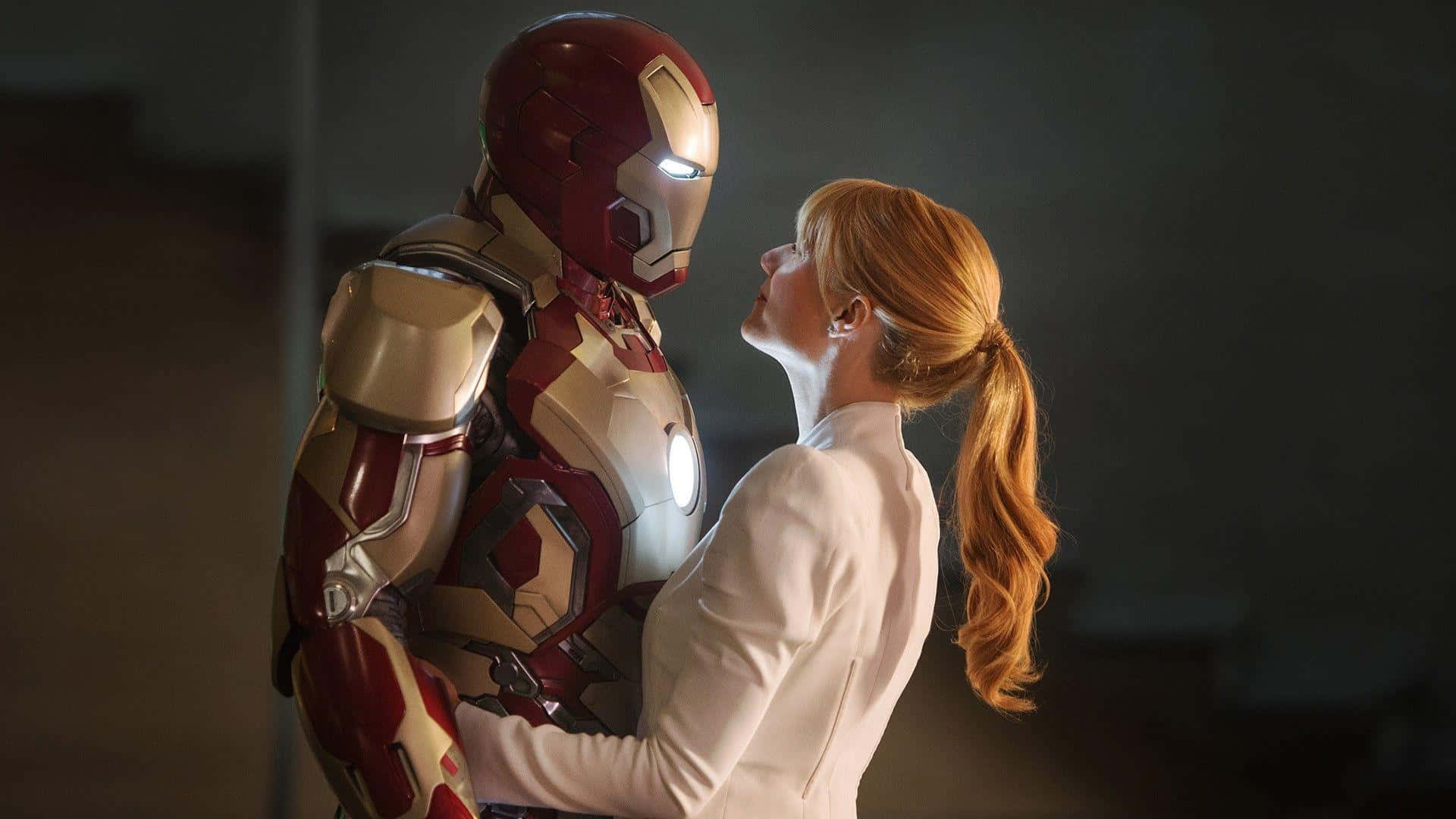 Pepper Potts 桌布 壁紙 — 一位身穿白色西裝的女性在昏暗的房間裡擁抱鐵甲奇俠，柔和的光線突顯了他們親密的時刻。