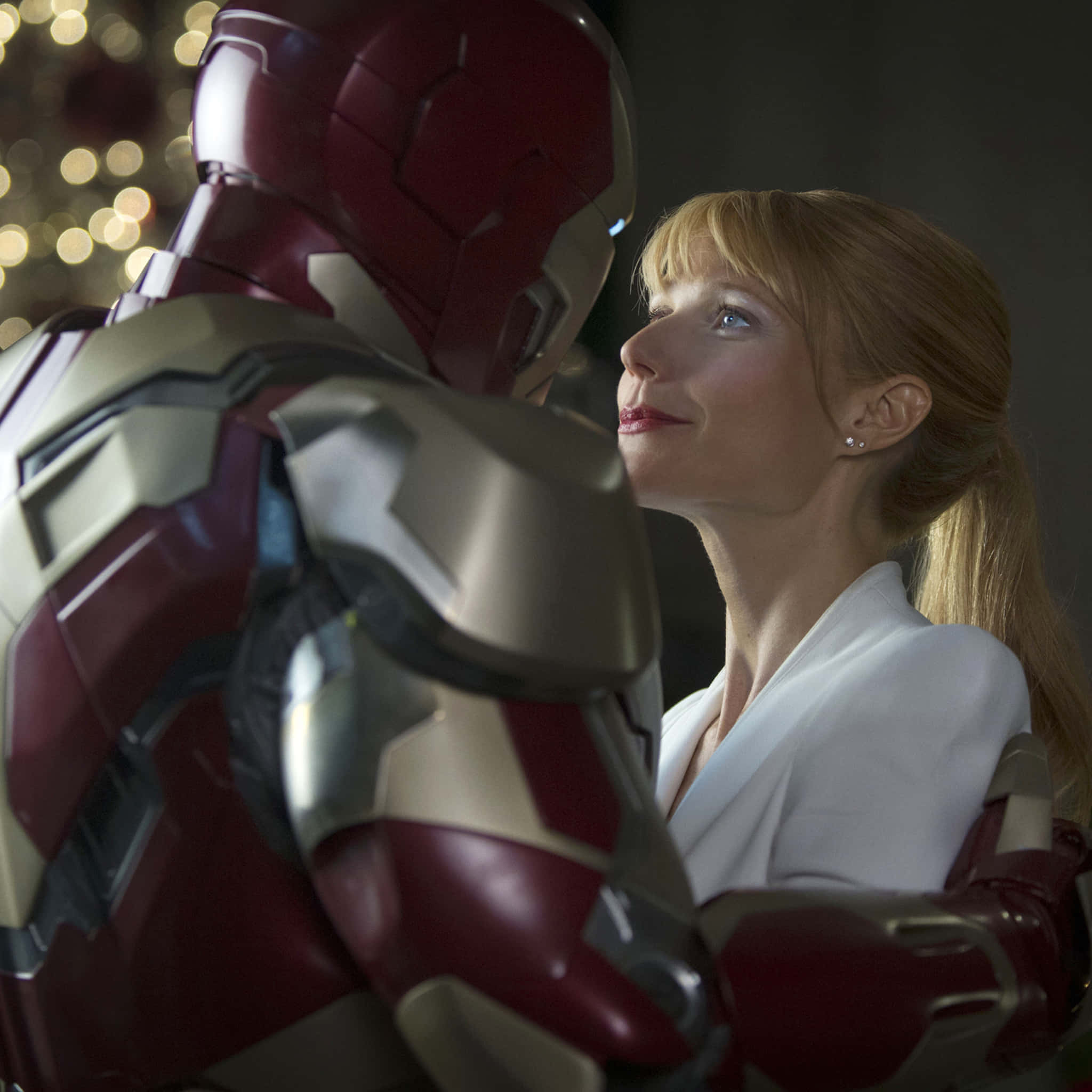 Pepper Potts 桌布 壁紙 — 一位身穿白色西裝的女性在溫暖的室內場景中，抬頭仰望鐵甲奇俠的紅色和金色裝甲，背景有散景燈光。