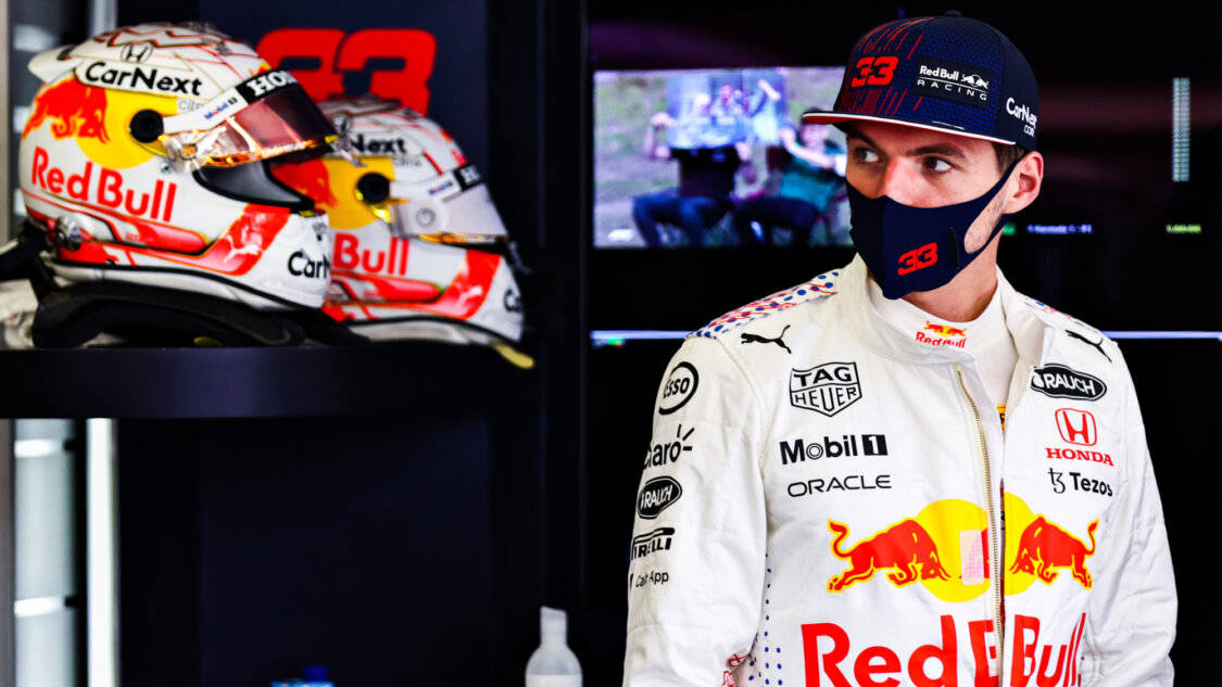 Verstappen 桌布 壁紙 — 一位身穿白色賽車服和帽子的紅牛車隊車手，在光線昏暗的車隊車庫中站在他的頭盔旁邊。