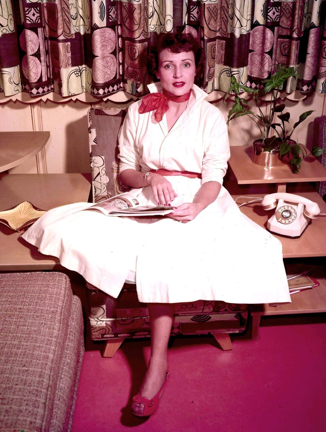 Betty White 桌布 壁紙 — 一位身穿白色連衣裙、帶有紅色點綴的女性，優雅地坐在一間擁有圖案窗簾和旋轉撥號電話的中世紀復古客廳中。