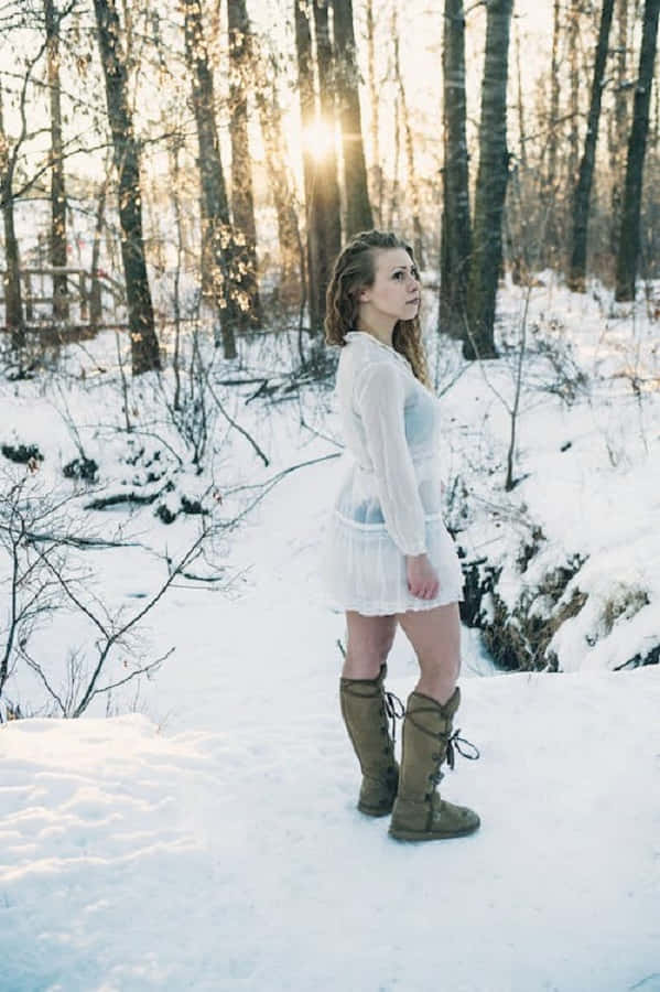 Snowshoes 桌布 壁紙 — 一位身穿白色連衣裙和棕色靴子的女士站在雪地森林中，金色的陽光穿過光禿禿的樹木灑落下來。