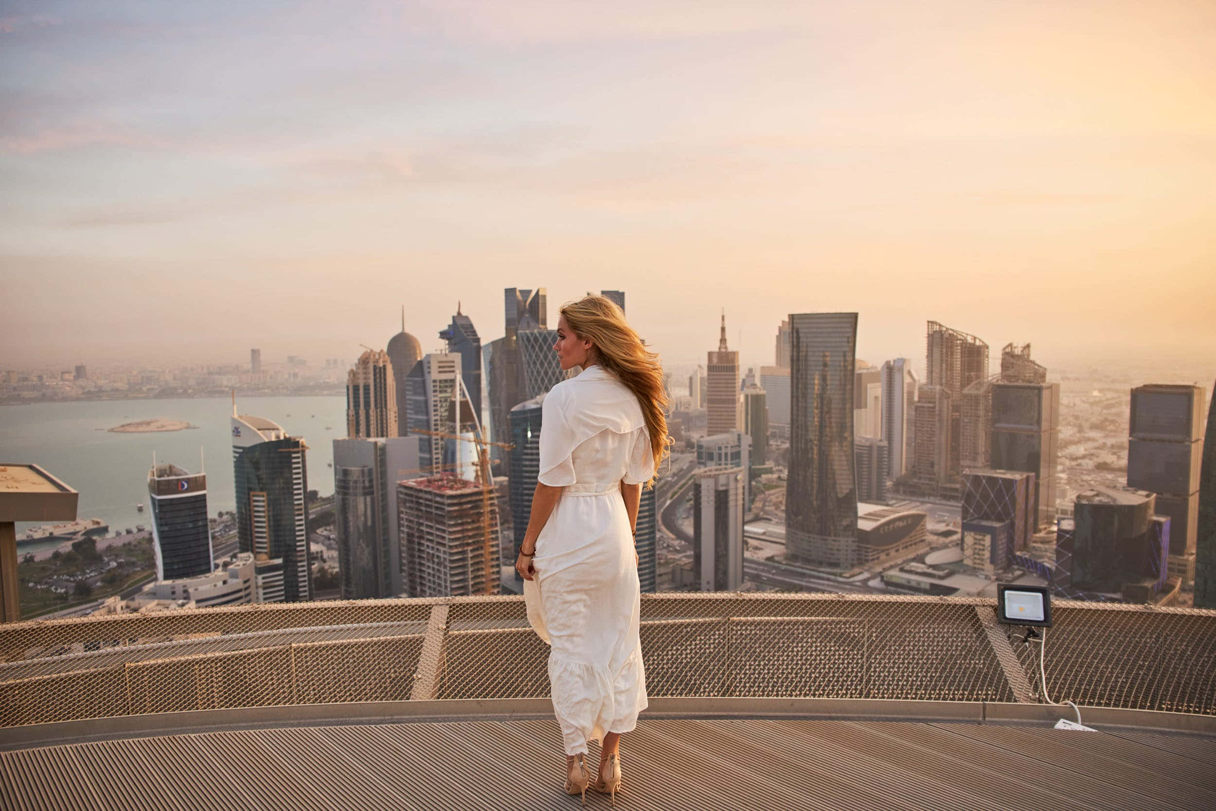 Qatar 桌布 壁紙 — 一位身穿白色連衣裙的女性站在屋頂上，俯瞰著現代城市的天際線，正值黃金時刻，光線溫暖。