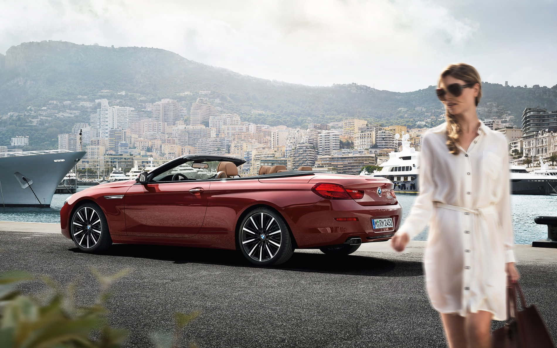 Bmw 6 Series 桌布 壁紙 — 一位身穿白色連衣裙的女性走過一輛停泊在港口的紅色敞篷車，背景是豪華遊艇和山巒環繞的城市景觀。