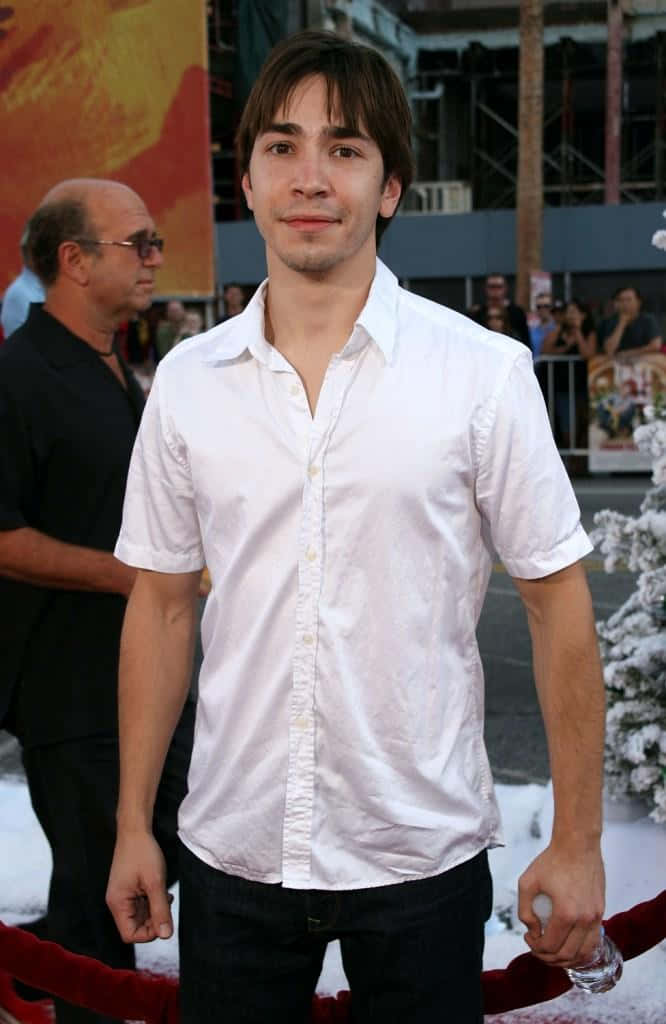 Justin Long 桌布 壁紙 — 一位身穿白色鈕扣襯衫的年輕男子站在紅地毯上，身後是積雪的樹木和活動人群。